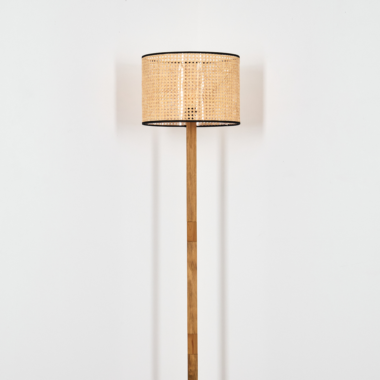 Eine Stehlampe mit gewebtem Lampenschirm und Holzständer. Der Lampenschirm ist beige mit schwarzem Rand. Der Hintergrund ist eine weiße Wand.