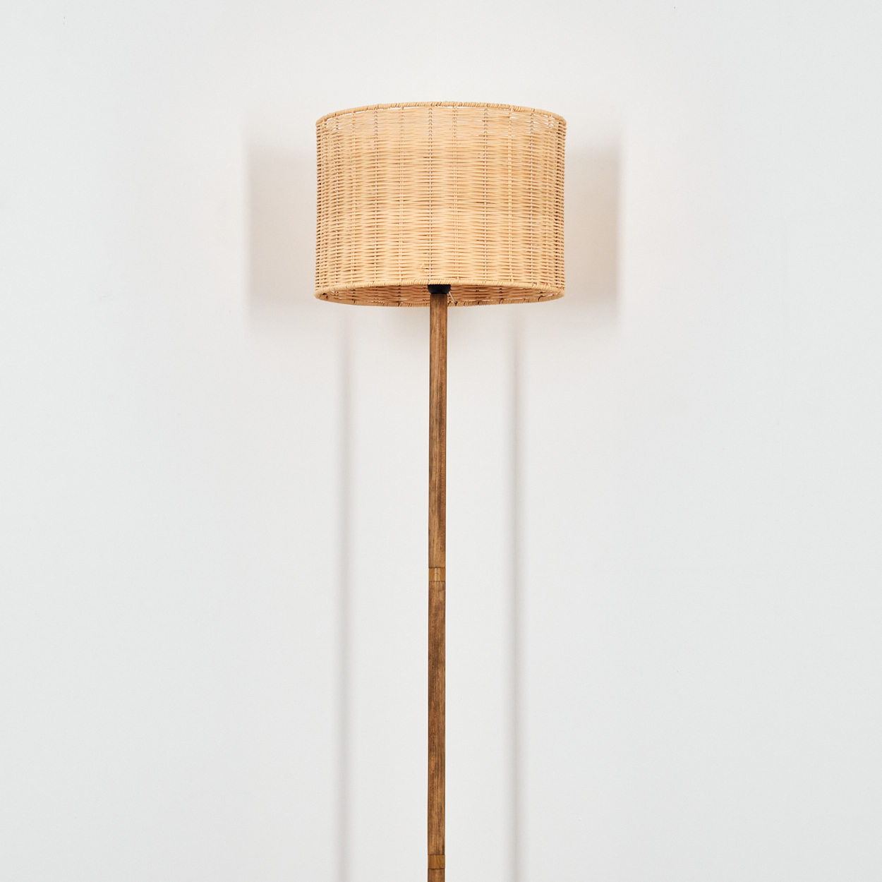 Eine Stehlampe mit gewebtem Lampenschirm steht vor einer weißen Wand. Der Lampenschirm ist beige, zylindrisch, und der Pfahl ist aus Holz.