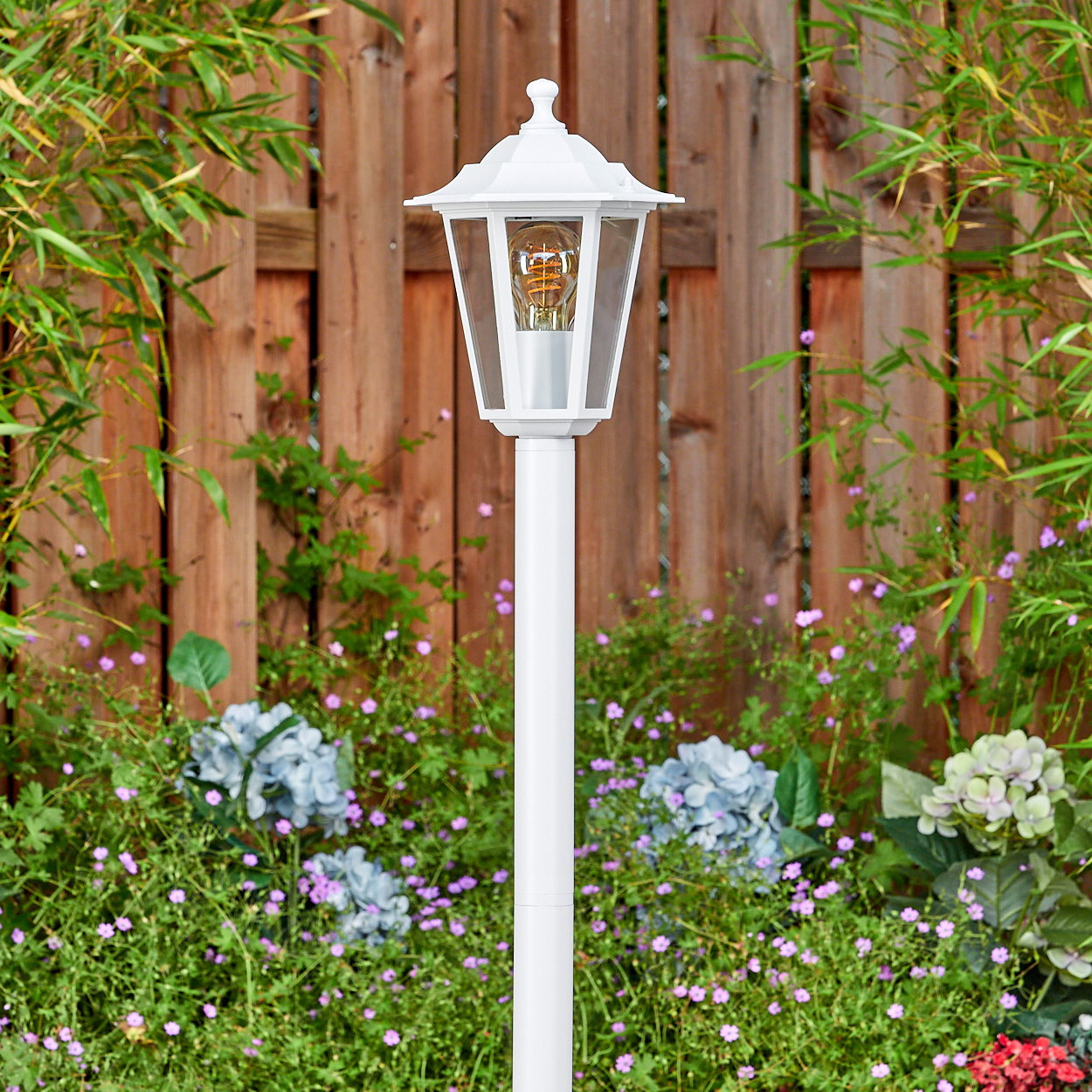 Weiße Außenlampe mit sichtbarer Glühbirne, in einem Garten aufgestellt. Im Hintergrund ein Holzzaun und grüne Pflanzen.