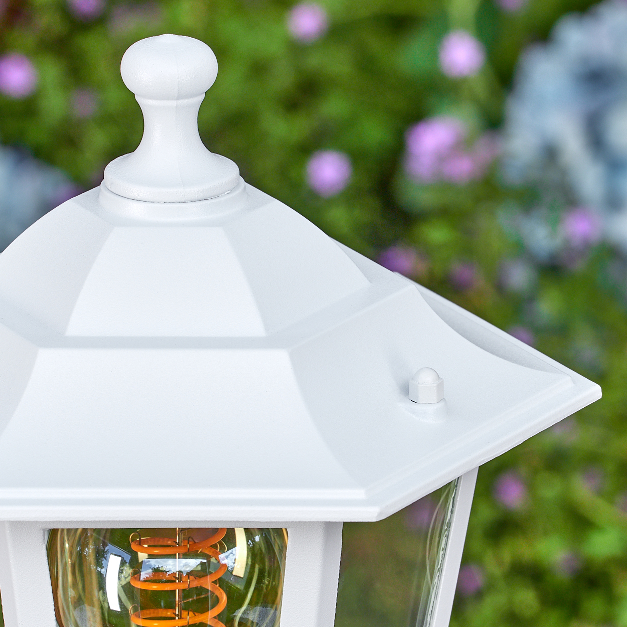 Nahaufnahme einer weißen Außenlampe. Die Lampe hat einen Glasteil mit einer warmen Glühbirne. Grünes Laub und rosa Blüten im Hintergrund.
