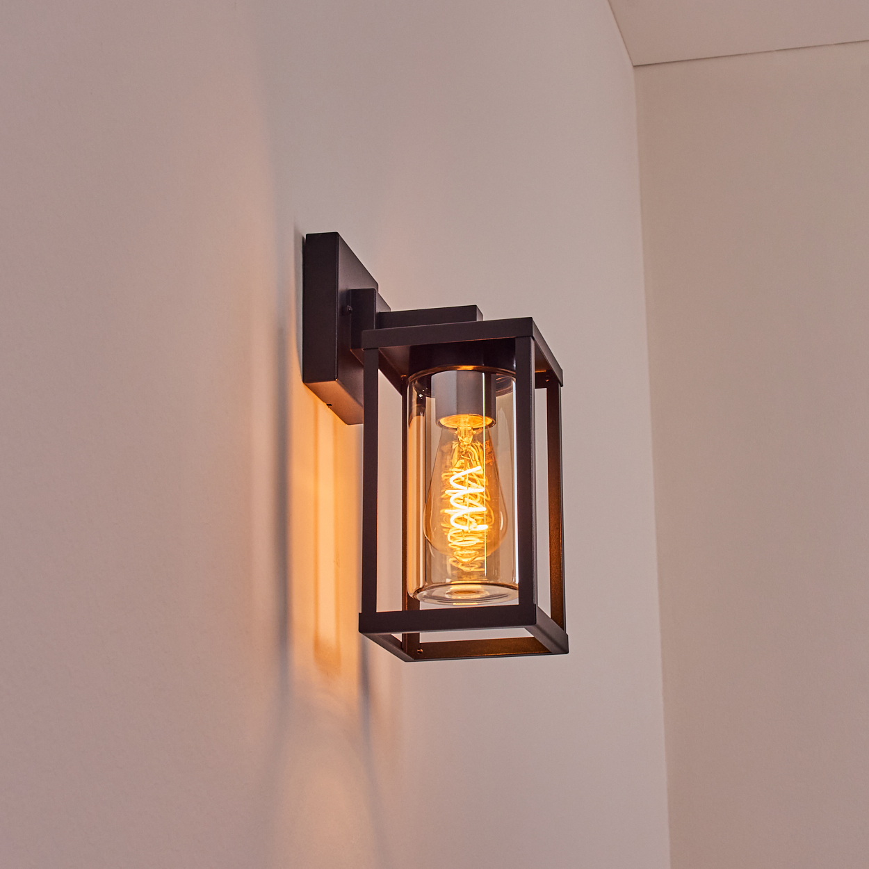 Eine schwarze Wandleuchte ist an einer weißen Wand montiert. Sie hat einen Glaszylinder mit einer gelb leuchtenden Glühbirne.