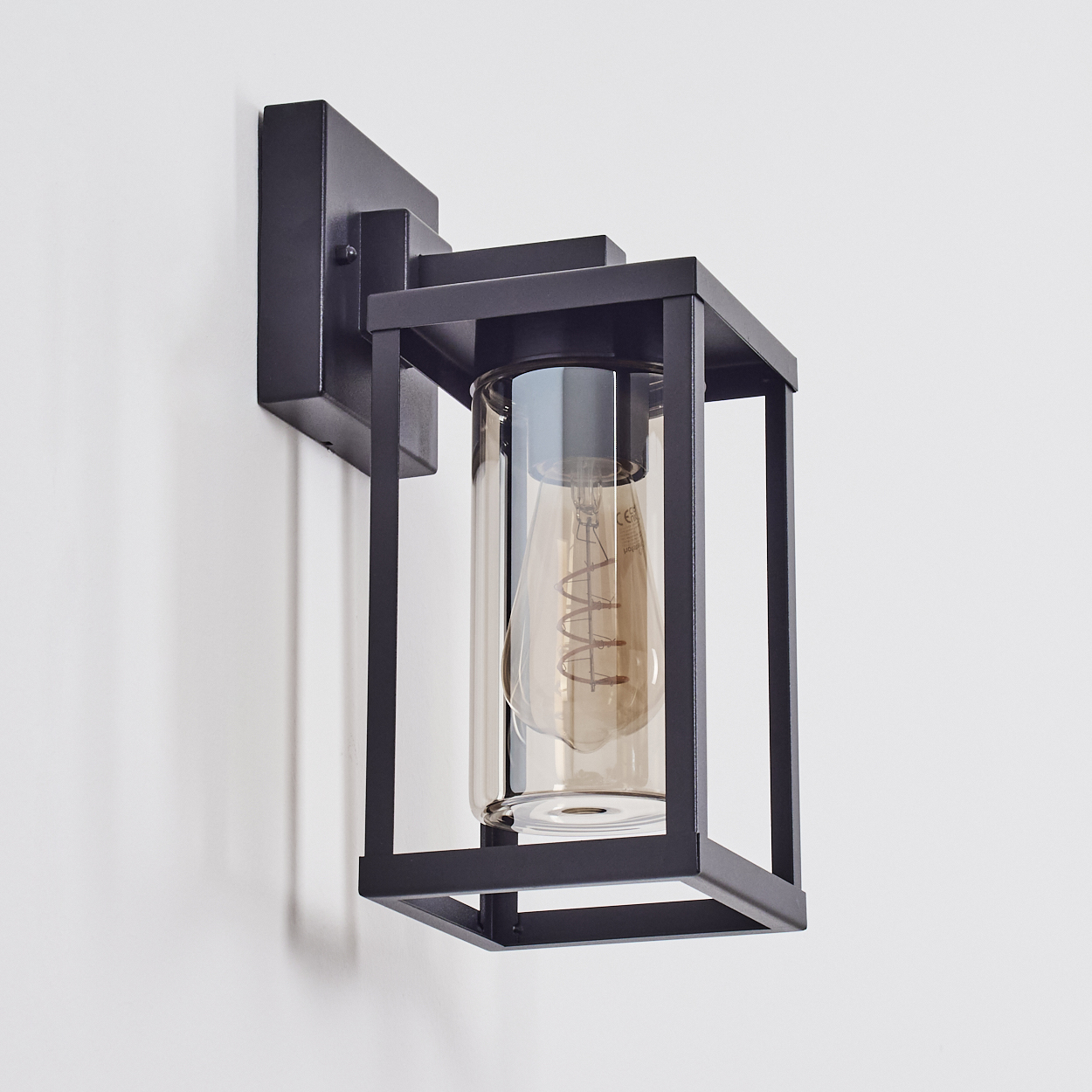 Schwarze Außenwandleuchte. Sie hat einen Glasschirm mit einer Glühbirne im Inneren, umgeben von einem schwarzen Metallrahmen. An einer weißen Wand montiert.