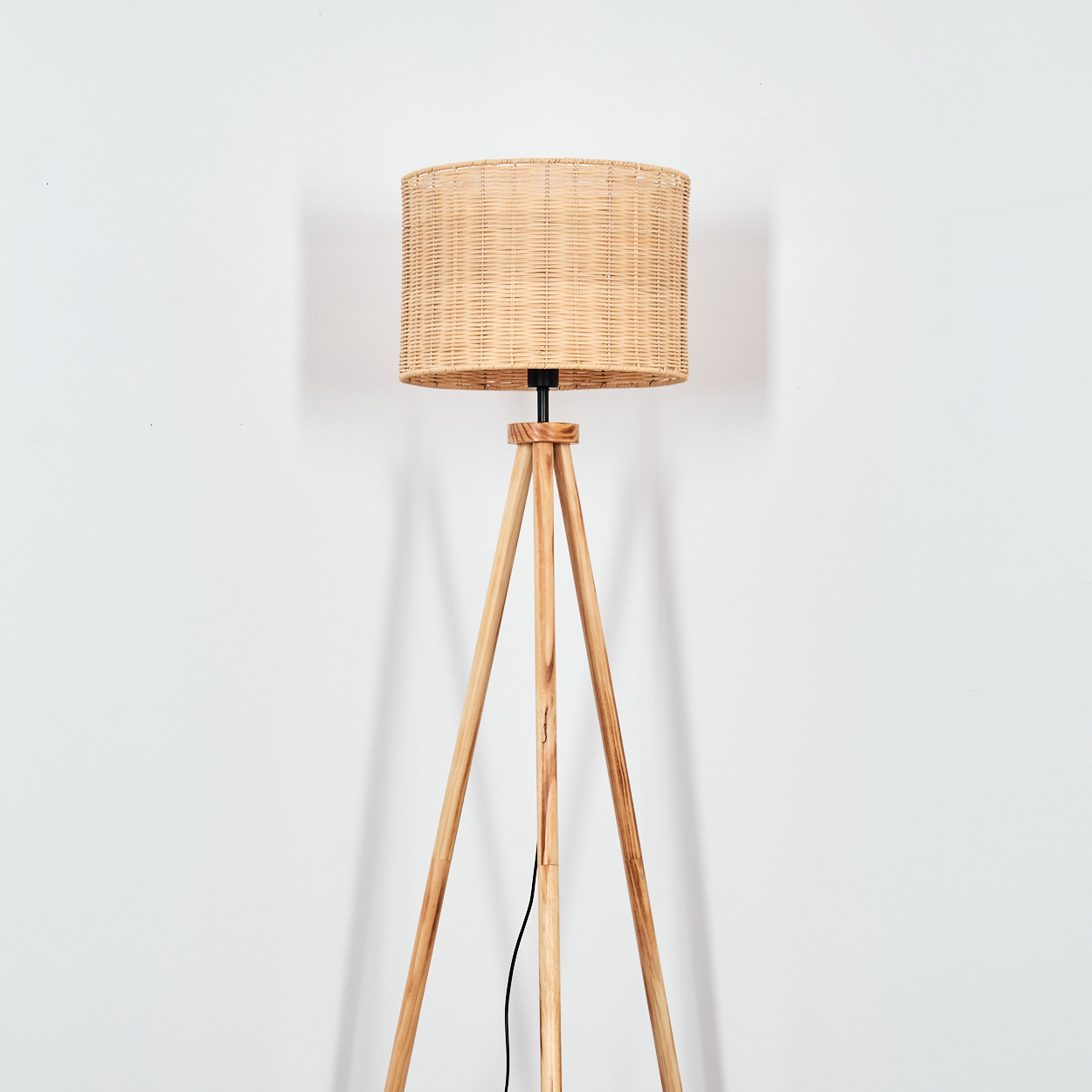 Eine hölzerne Stativ-Stehlampe mit gewebtem Lampenschirm steht vor einer weißen Wand. Ein schwarzes Kabel verläuft am Bein der Lampe.