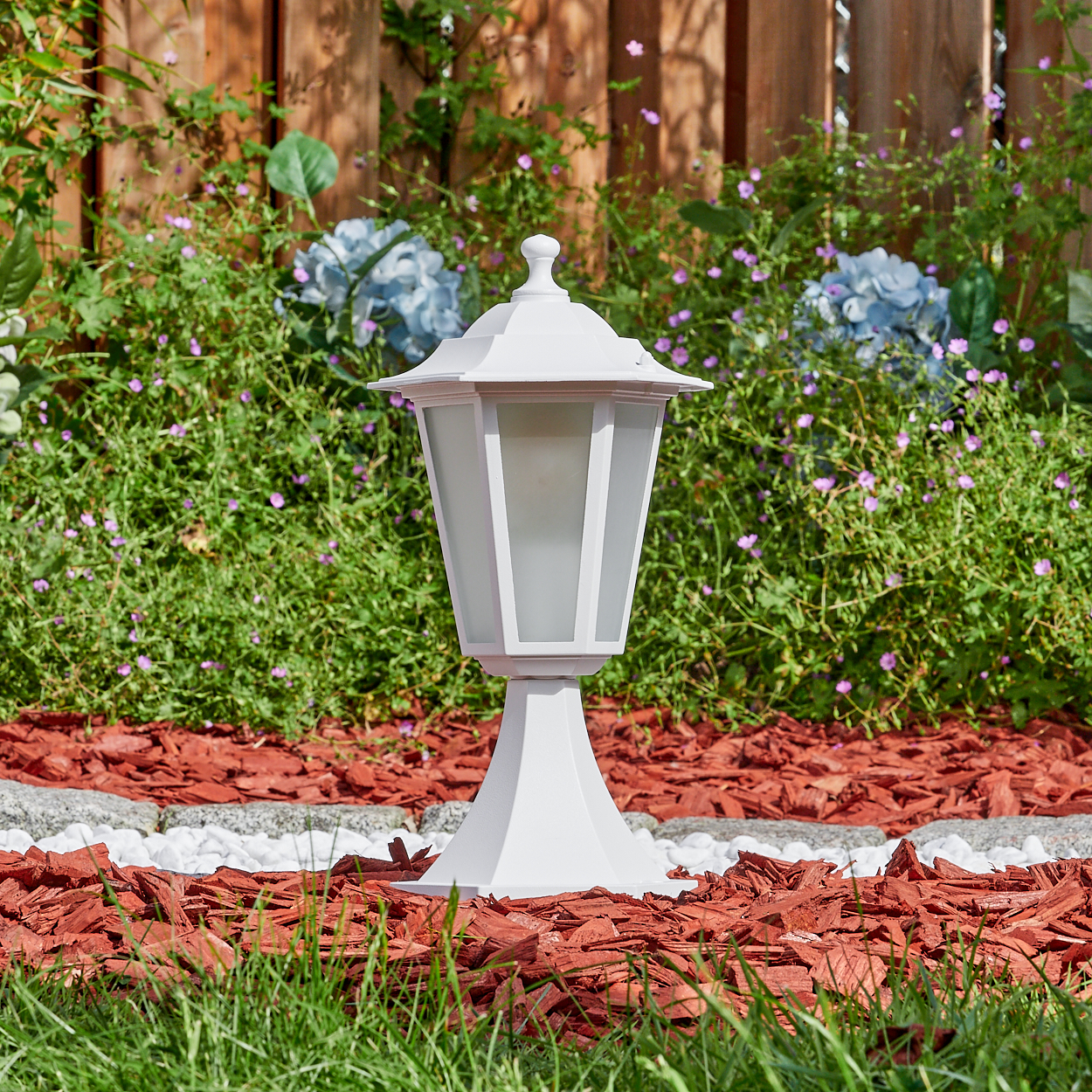 Eine weiße Außenlampe steht in einem Garten mit roter Mulch, weißen Steinen und grünem Gras. Blaue Blumen und ein Holzzaun sind im Hintergrund.