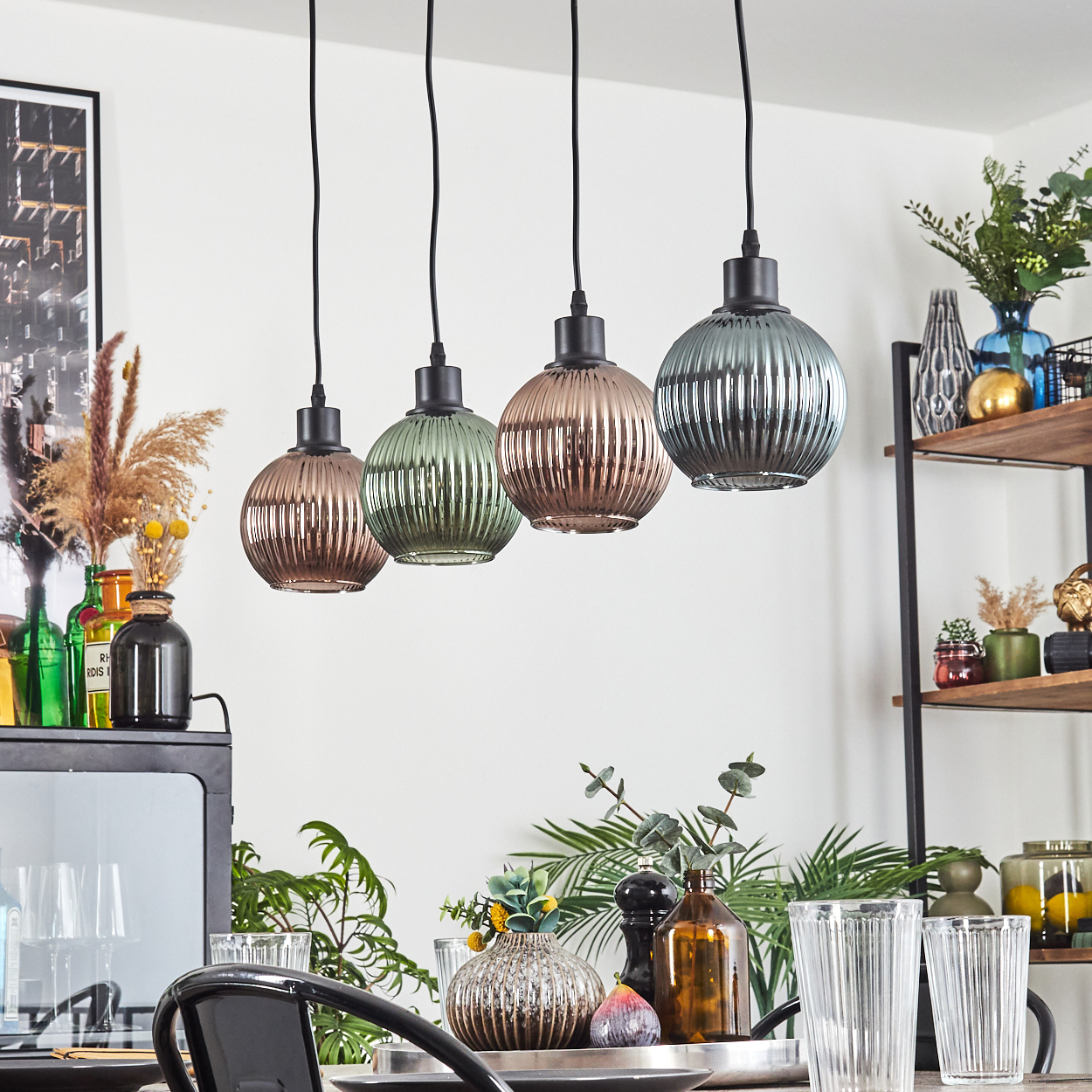 Vier Glas-Pendelleuchten hängen über einem Tisch mit Gläsern und Dekoration. Regale mit Pflanzen und Dekoration sind im Hintergrund sichtbar.