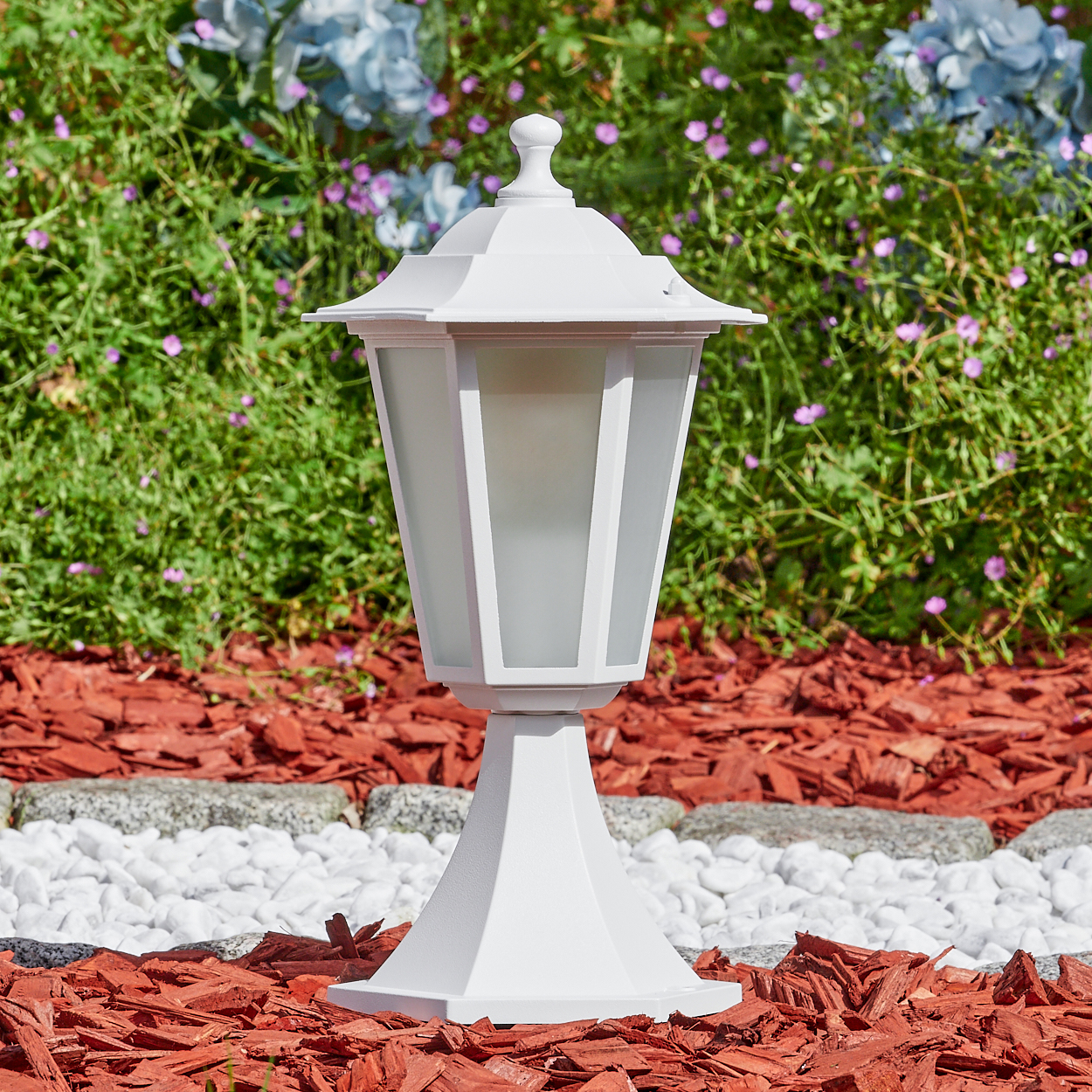 Eine weiße Außenlampe steht auf Mulch. Dahinter befindet sich ein grüner Busch mit blauen und lila Blüten.