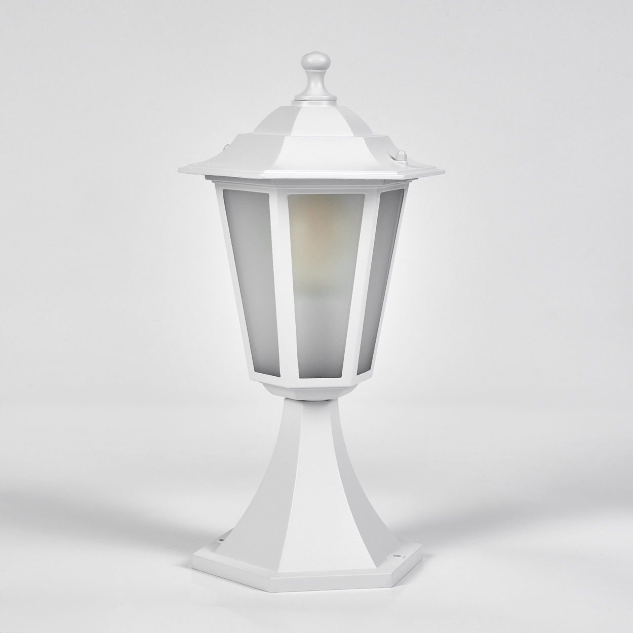 Eine weiße Außenlampe steht auf einer weißen Oberfläche. Sie hat einen sechseckigen Körper mit Glasscheiben und eine spitze Spitze.