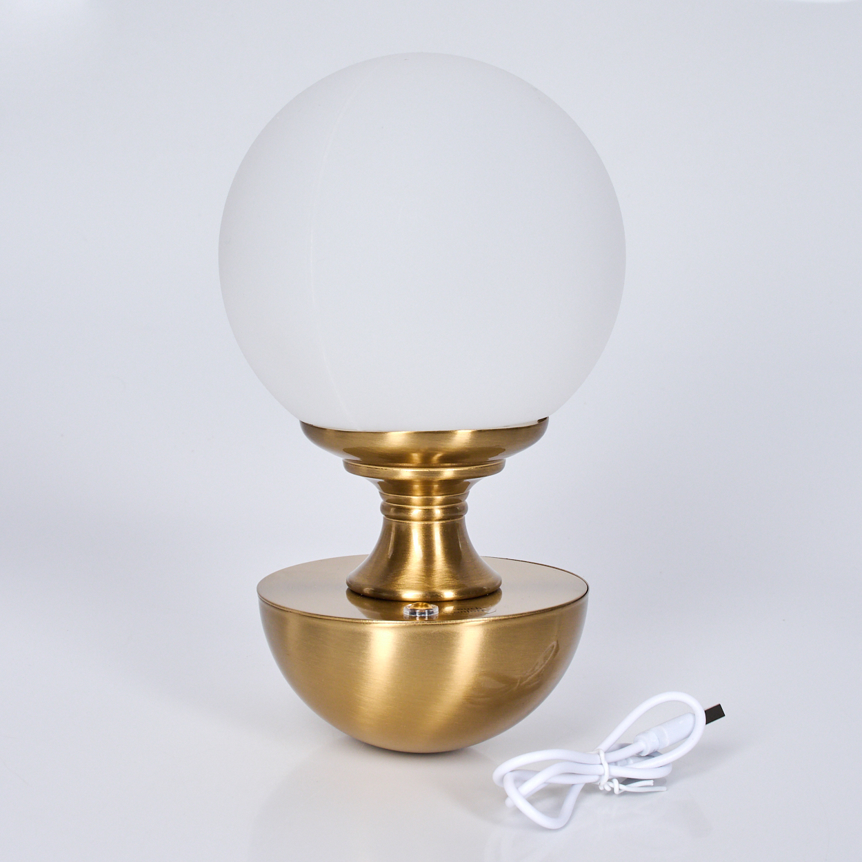 Eine goldene Tischlampe mit weißem Kugelschirm steht auf einer weißen Oberfläche. Rechts befindet sich ein weißes USB-Kabel.