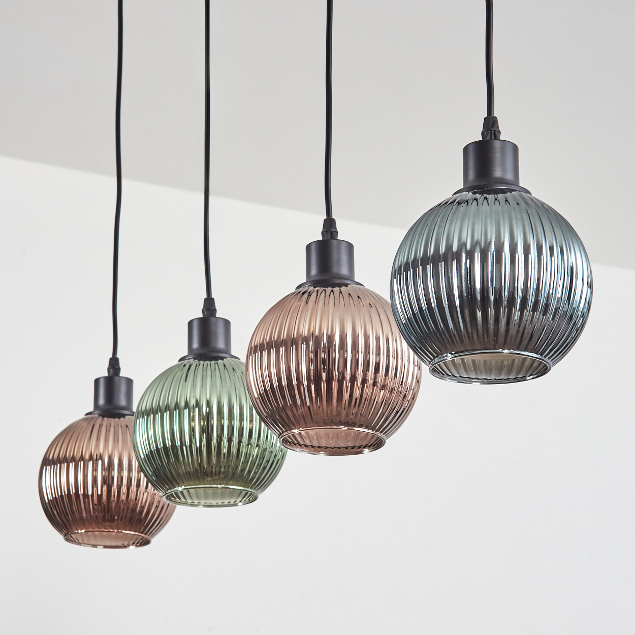 Vier Pendelleuchten mit gerippten Glasschirmen in Kupfer-, Grün- und Grautönen, die an schwarzen Kabeln von einer weißen Decke hängen.
