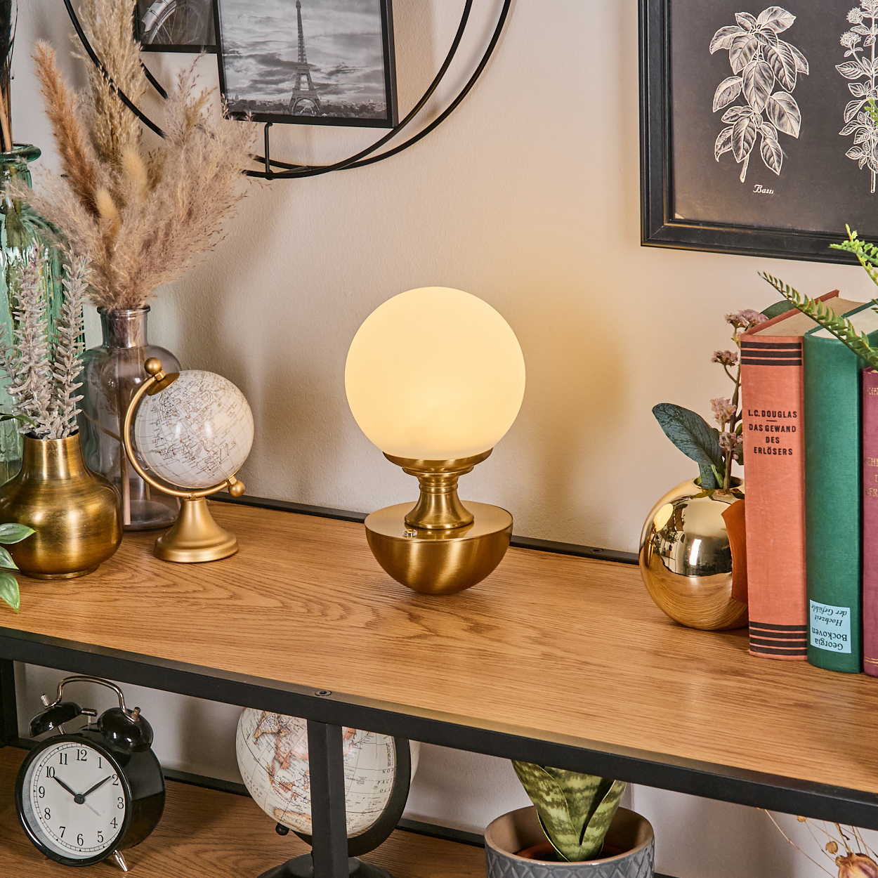 Ein Regal mit Lampe, Büchern, Globen und Dekorationen. Auf dem unteren Regal befindet sich ein schwarzer Wecker. Im Hintergrund ist ein Bild des Eiffelturms zu sehen.