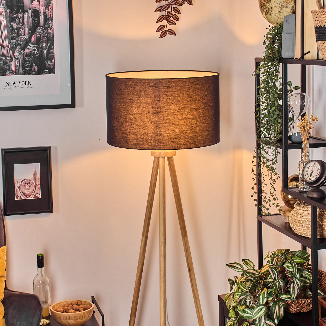 Eine Stehlampe mit Holzstativ und dunklem Lampenschirm, neben einem Regal mit Pflanzen und Dekoration sowie gerahmten Bildern an der Wand.
