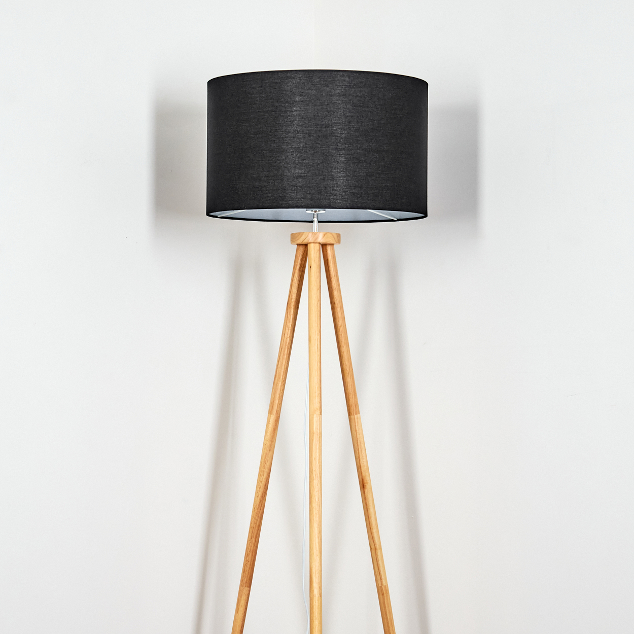 Eine Stehlampe mit schwarzem Lampenschirm und hölzernen Stativbeinen steht vor einer weißen Wand.