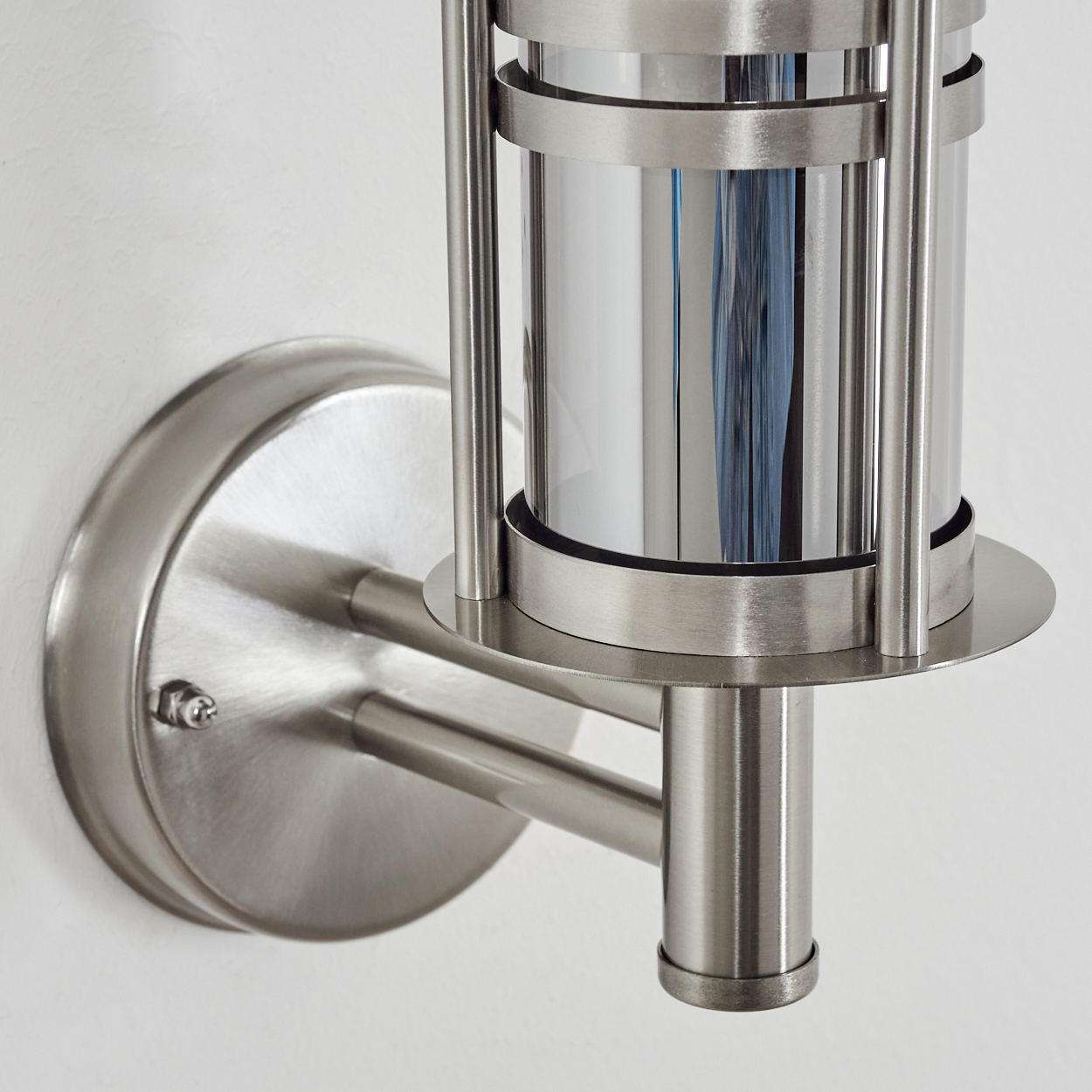 Eine metallische Wandleuchte mit zylindrischer Lampe. Sie hat eine gebürstete Stahloberfläche und ist an einer weißen Wand montiert.