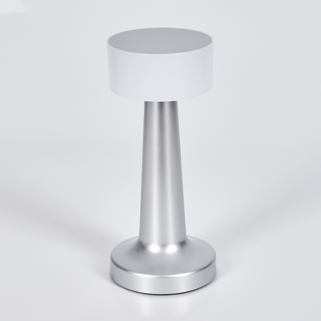 Eine silberne Tischlampe mit weißem Lampenschirm. Die Lampe hat einen zylindrischen Sockel, einen konischen Körper und einen runden Schirm.