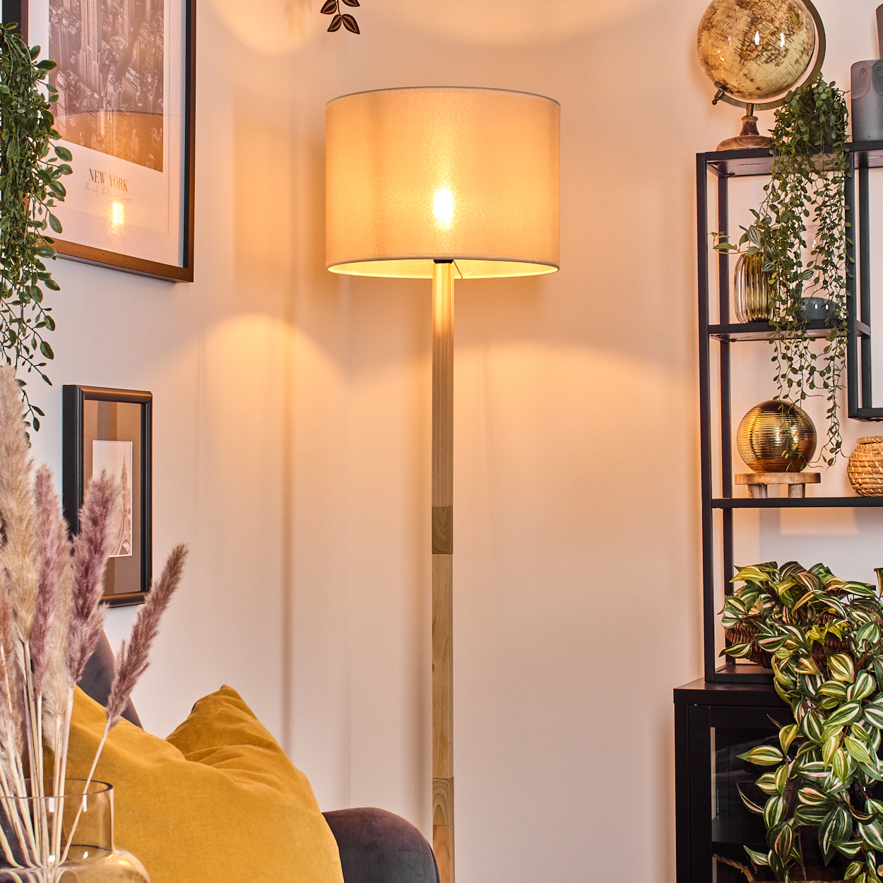 Eine beleuchtete Stehlampe mit beigem Schirm steht in einem Raum. Rechts ein Regal mit Pflanzen und einem Globus. Ein Bild hängt an der Wand.