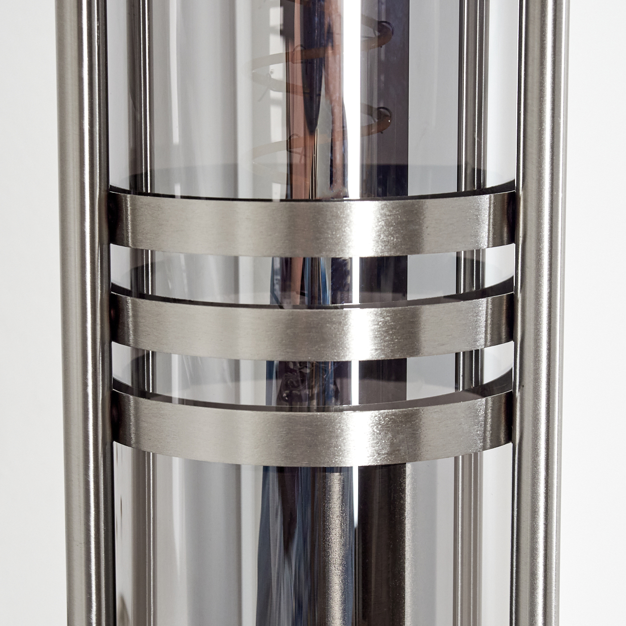 Nahaufnahme einer modernen Lampe. Sie hat einen zylindrischen Glaskern, umgeben von Metallstangen und horizontalen Bändern. Der Hintergrund ist weiß.