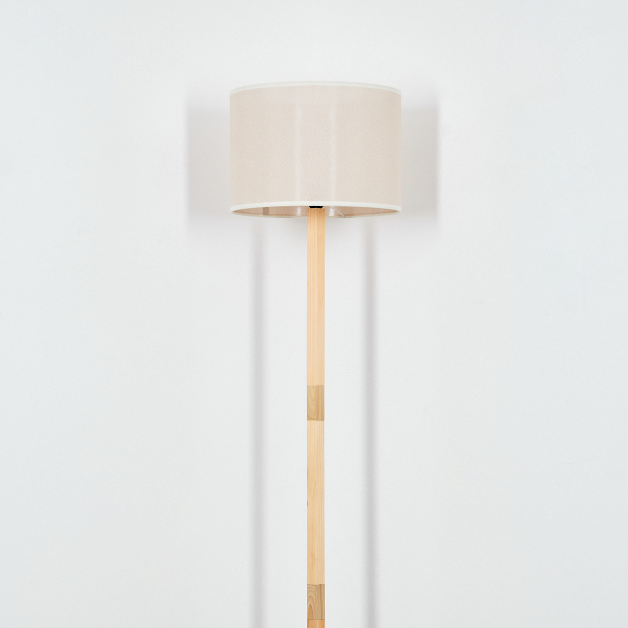 Ein beigefarbener Lampenschirm sitzt auf einem hellen Holzpfahl. Die Lampe steht vor einer weißen Wand und wirft einen Schatten.