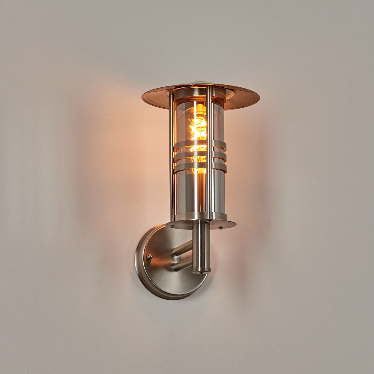 Eine Wandlampe mit zylindrischem Glasteil und einer brennenden Glühbirne. Sie hat eine metallische Oberfläche und ist an einer weißen Wand befestigt.