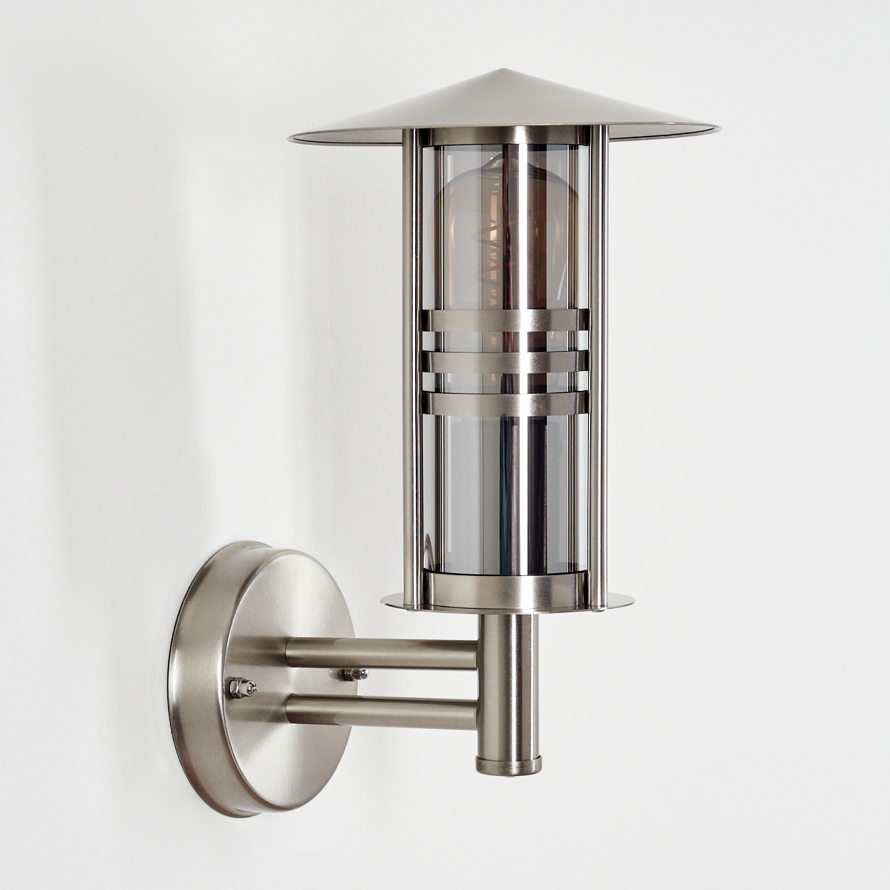 Eine moderne, silberne Wandlampe mit zylindrischem Glasschirm und konischem Deckel, an einer weißen Wand befestigt.