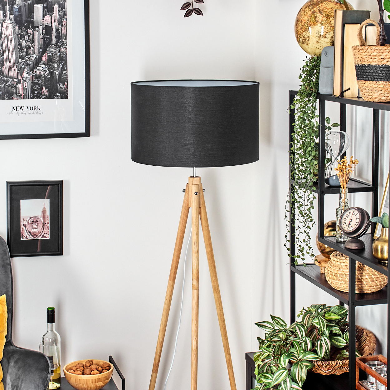 Eine schwarze Lampe mit Holzbeinen steht in der Nähe eines Regals mit Pflanzen und Dekorationen. Gemälde und eine Schüssel mit Nüssen sind in der Nähe.