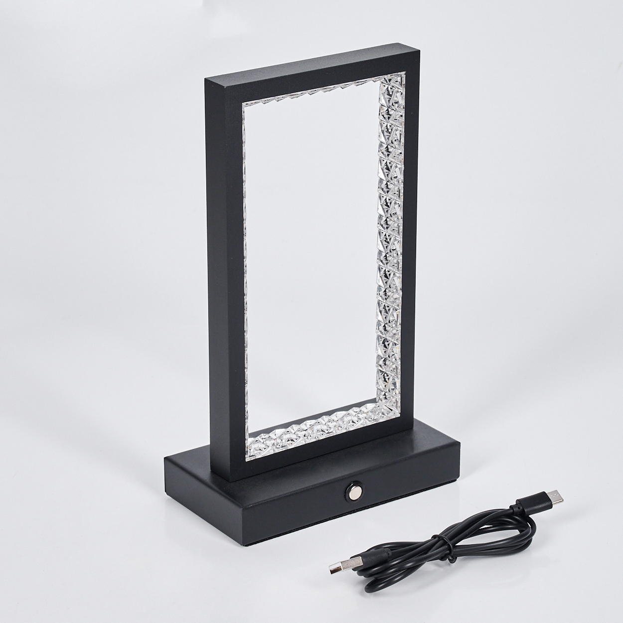 Eine schwarze rechteckige Lampe mit kristallartigen Kanten, auf einem Sockel, mit einem USB-Kabel. Vor weißem Hintergrund.