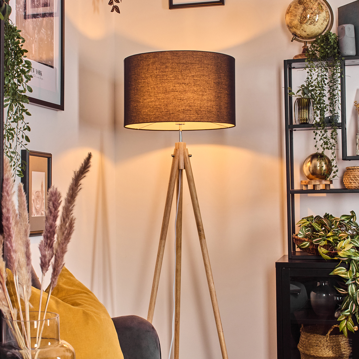 Eine Stehlampe mit Holzstativ und dunklem Schirm steht in einem Raum mit Pflanzen und Bildern.