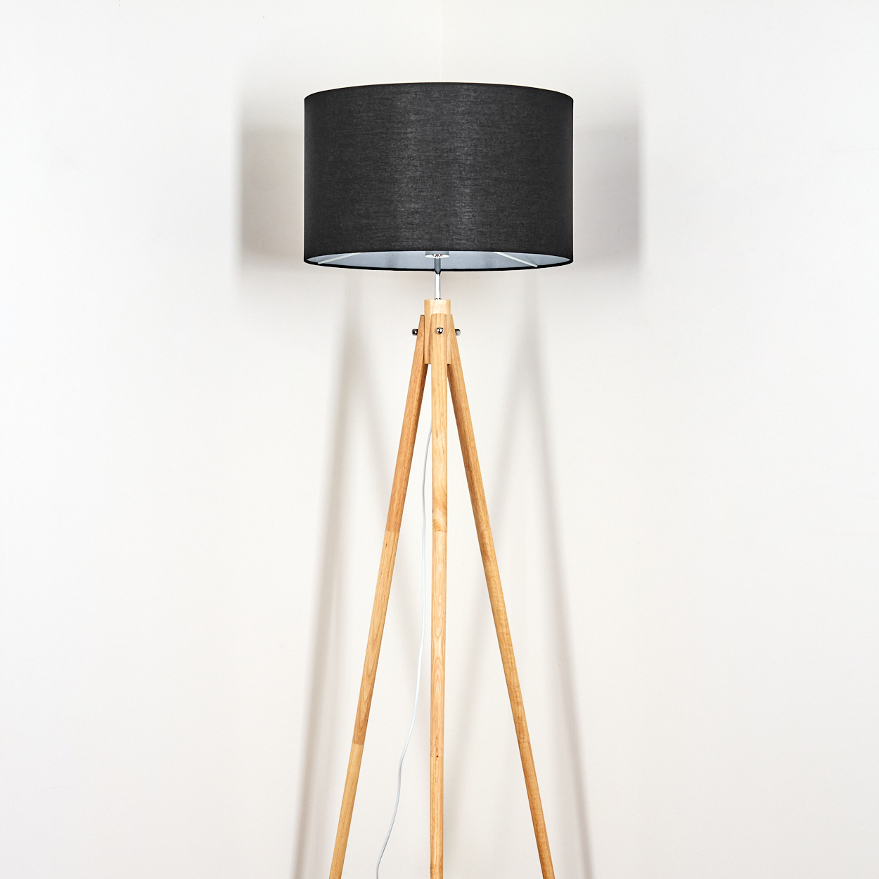 Eine Stehlampe aus Holz mit schwarzem Lampenschirm steht vor einer weißen Wand. Ein weißes Kabel verläuft ein Bein hinunter.
