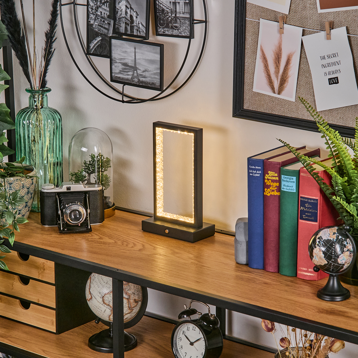 Holztisch mit Lampe, Büchern, Kamera, Uhr, Globus, Fotos und Pflanzen. Die Lampe hat ein leuchtendes Rechteck in der Mitte.