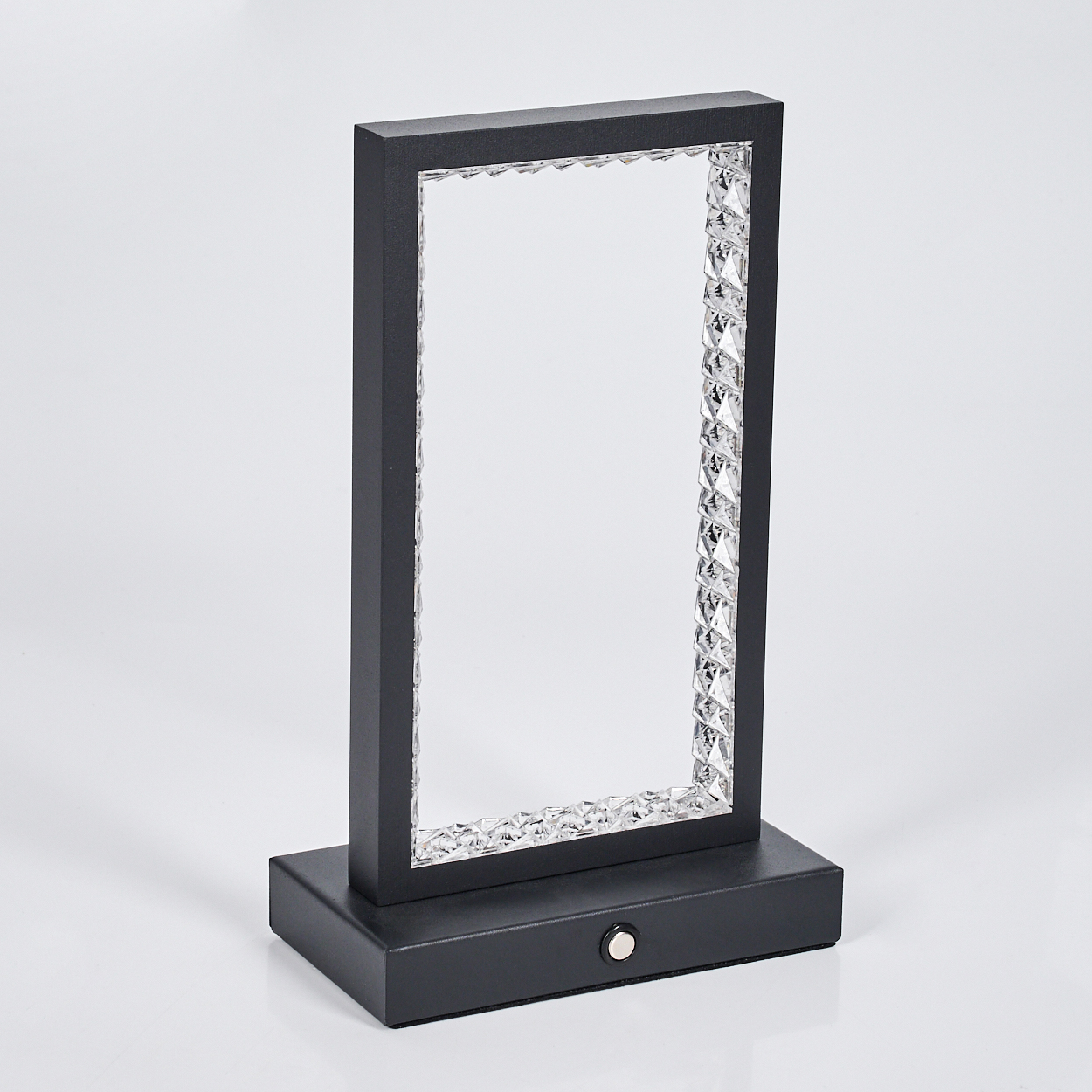 Eine schwarze rechteckige Lampe mit einem Rahmen aus Kristallen. Sie steht auf einem schwarzen Sockel mit einem kleinen Knopf. Der Hintergrund ist weiß.