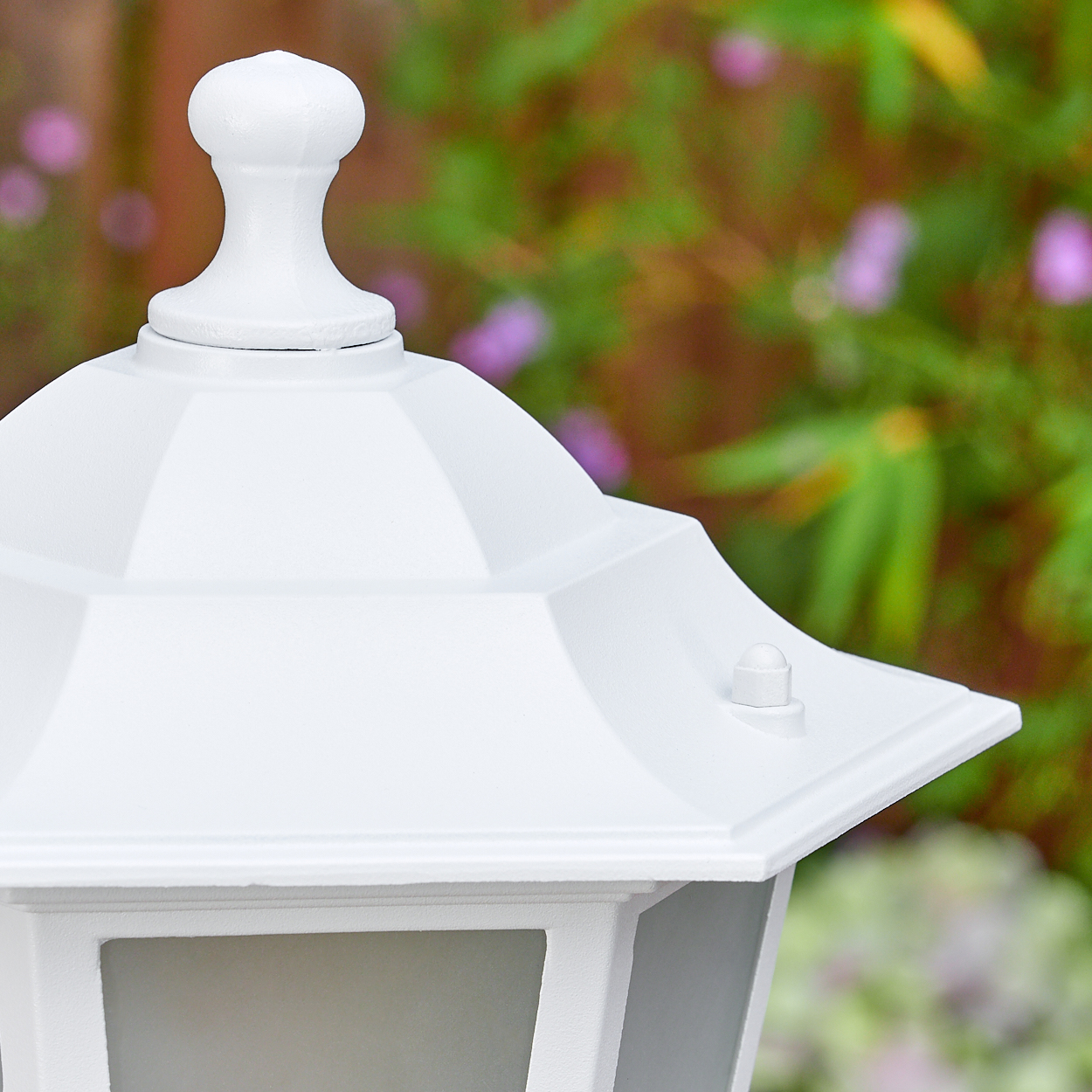 Nahaufnahme einer weißen Außenlampe mit dekorativer Spitze. Grünes Laub und Blumen sind im Hintergrund unscharf.