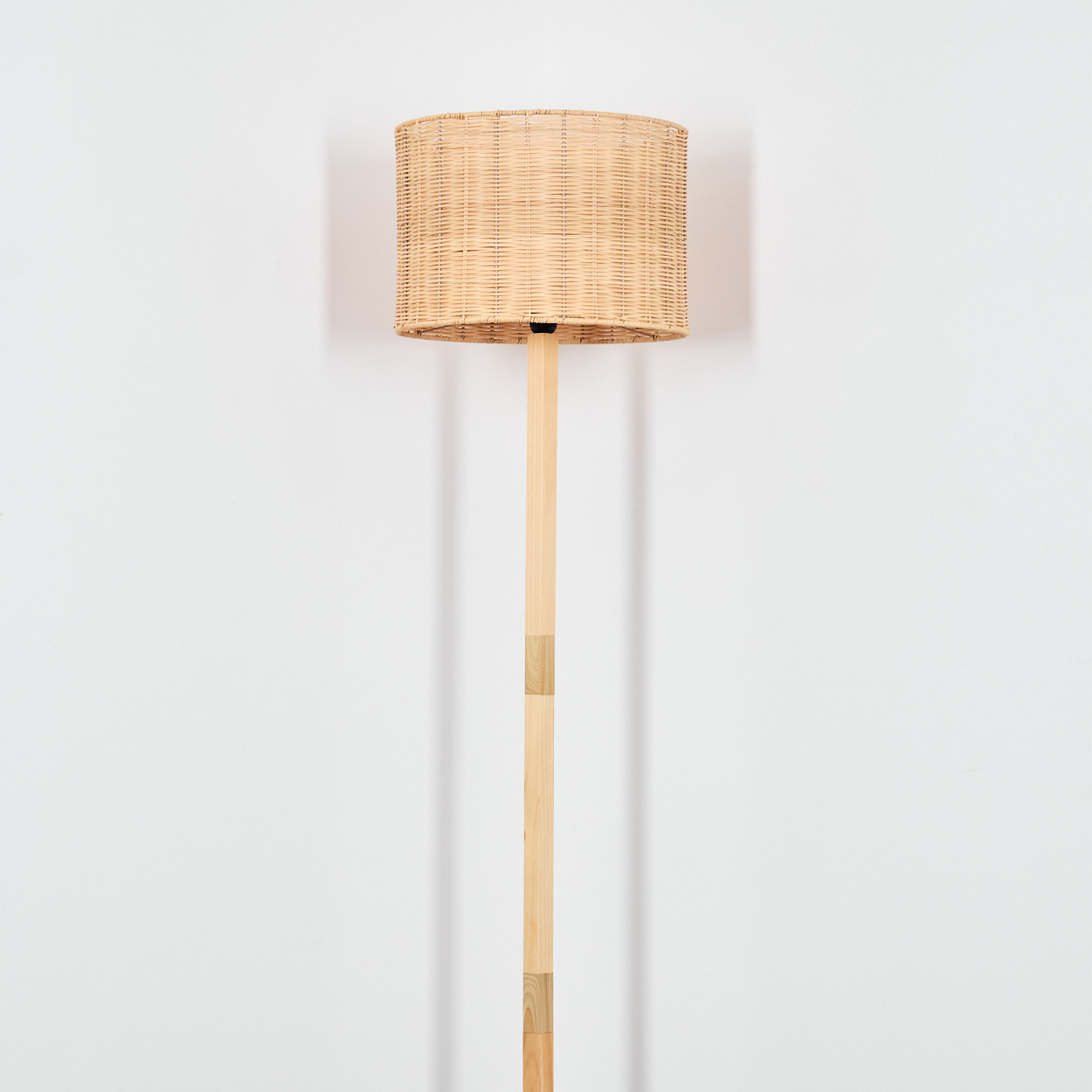 Eine Stehlampe mit gewebtem Lampenschirm und Holzstange steht vor einer weißen Wand.