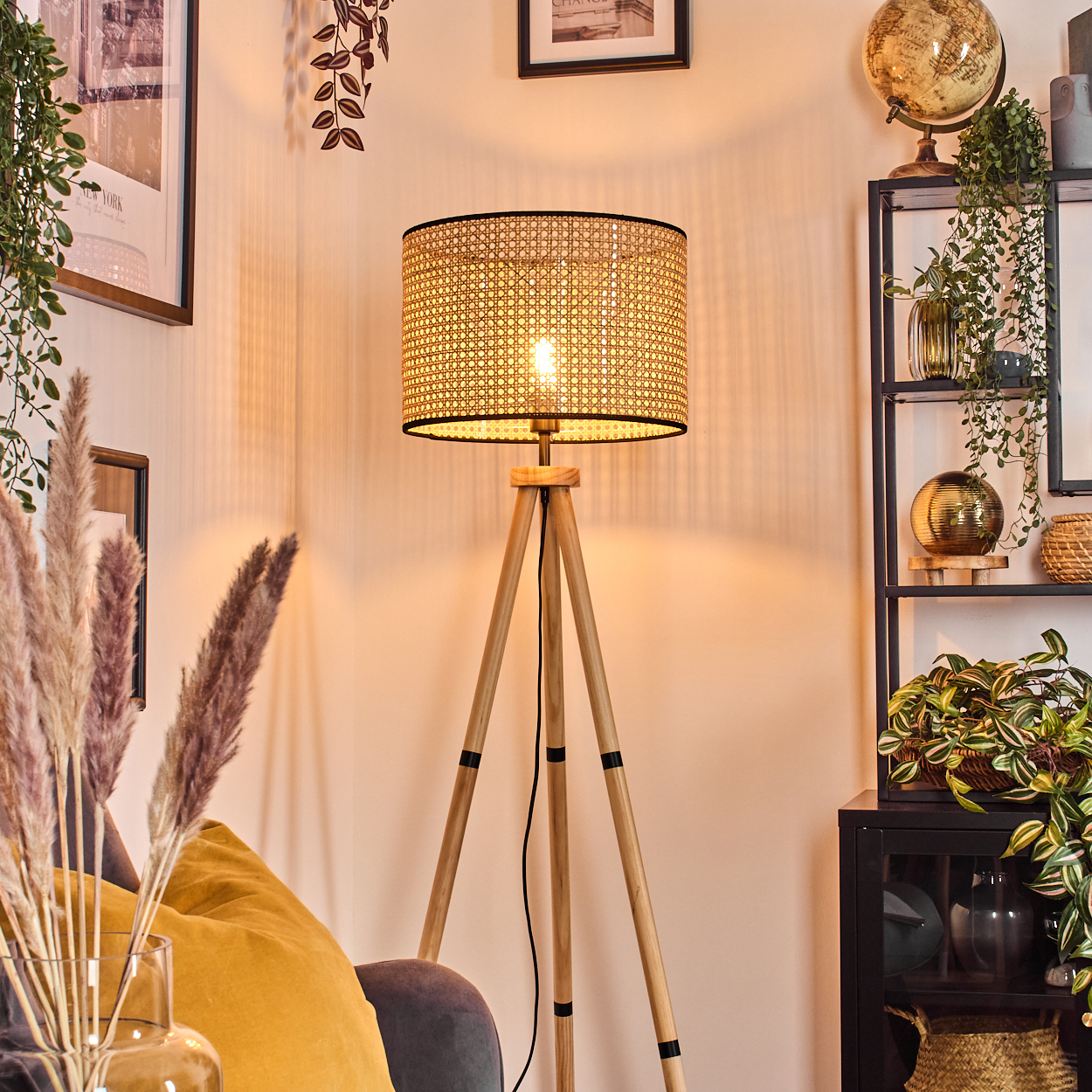 Eine beleuchtete Dreibein-Lampe mit gewebtem Schirm steht in einem Raum. Ein gelbes Kissen, Pampasgras und ein Bücherregal sind ebenfalls sichtbar.