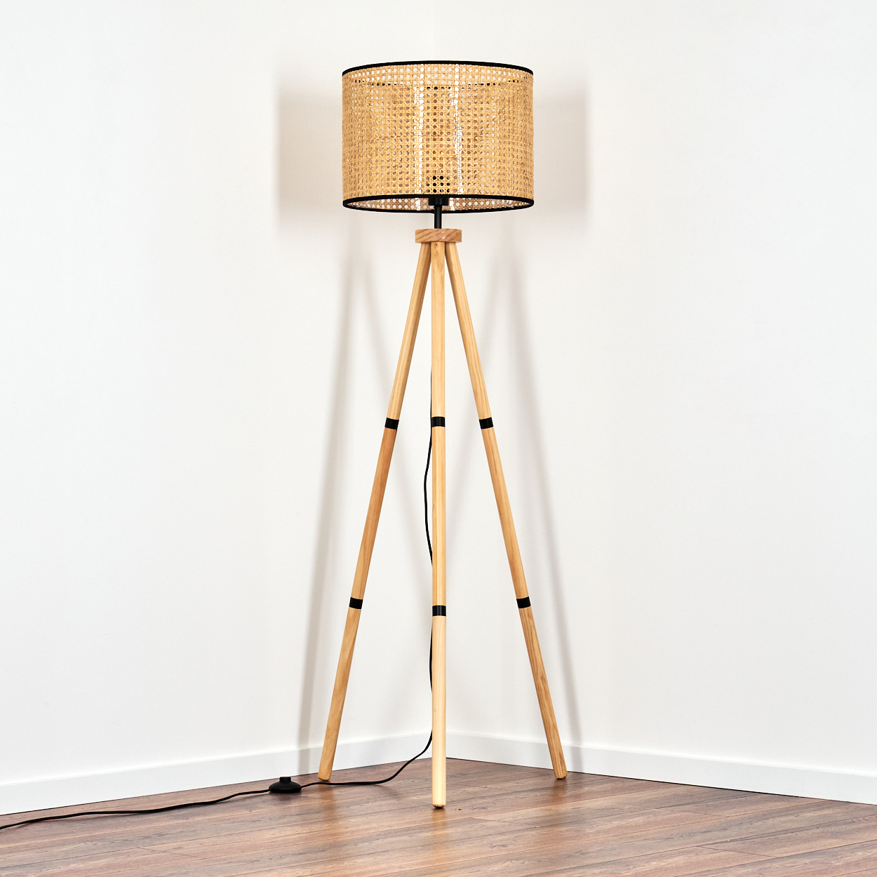 Eine hölzerne Stehlampe mit einem gewebten Schirm steht in einer Raumecke. Der Boden ist aus Holz und die Wände sind weiß.