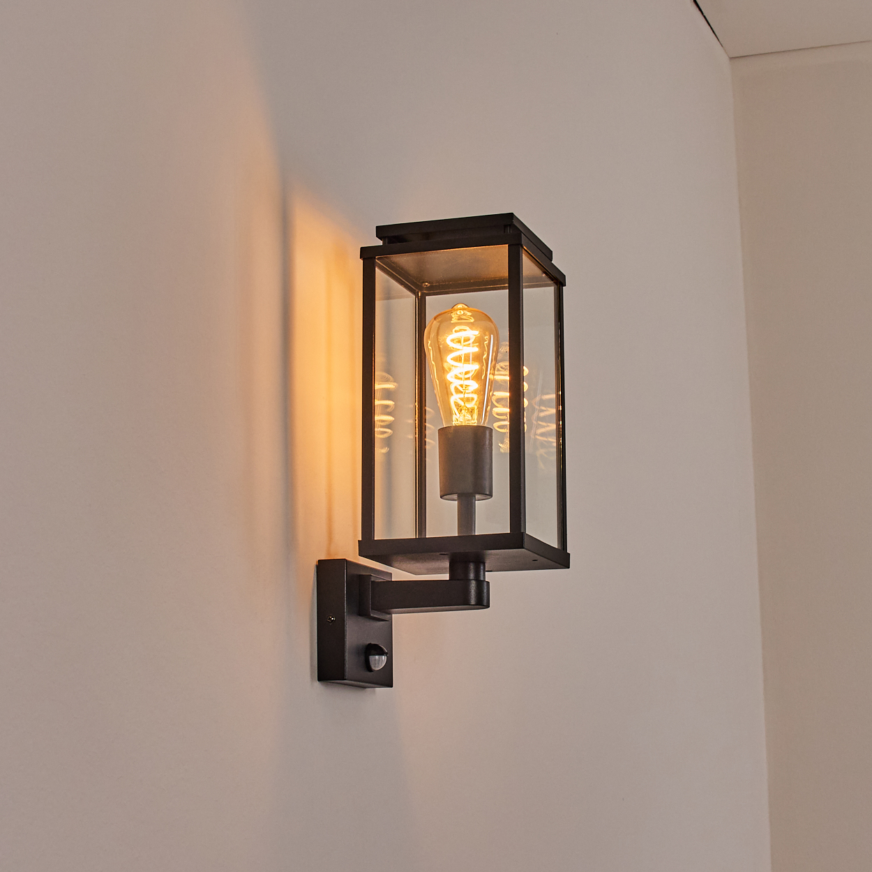 Eine schwarze Wandlampe mit rechteckigem Glasschirm. Im Inneren leuchtet eine orangefarbene Glühbirne. Die Lampe ist an einer weißen Wand montiert.