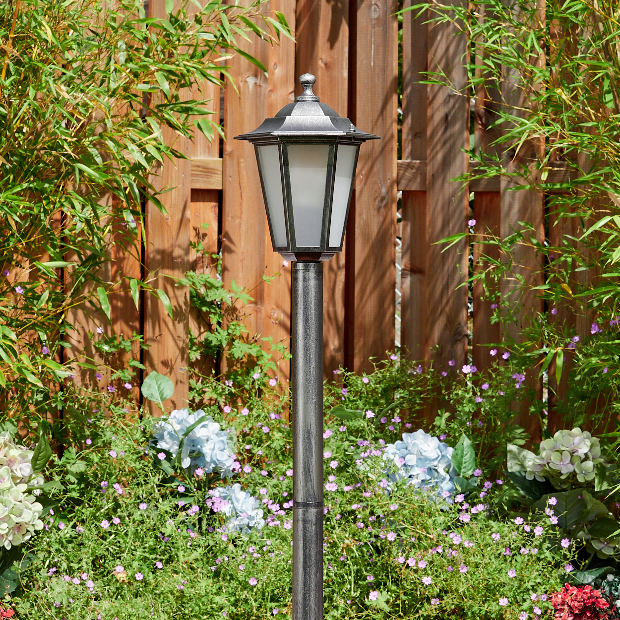 Eine metallische Gartenlampe steht vor einem Holzzaun. Grüne Pflanzen und Blumen umgeben die Lampe und schaffen eine natürliche Umgebung.