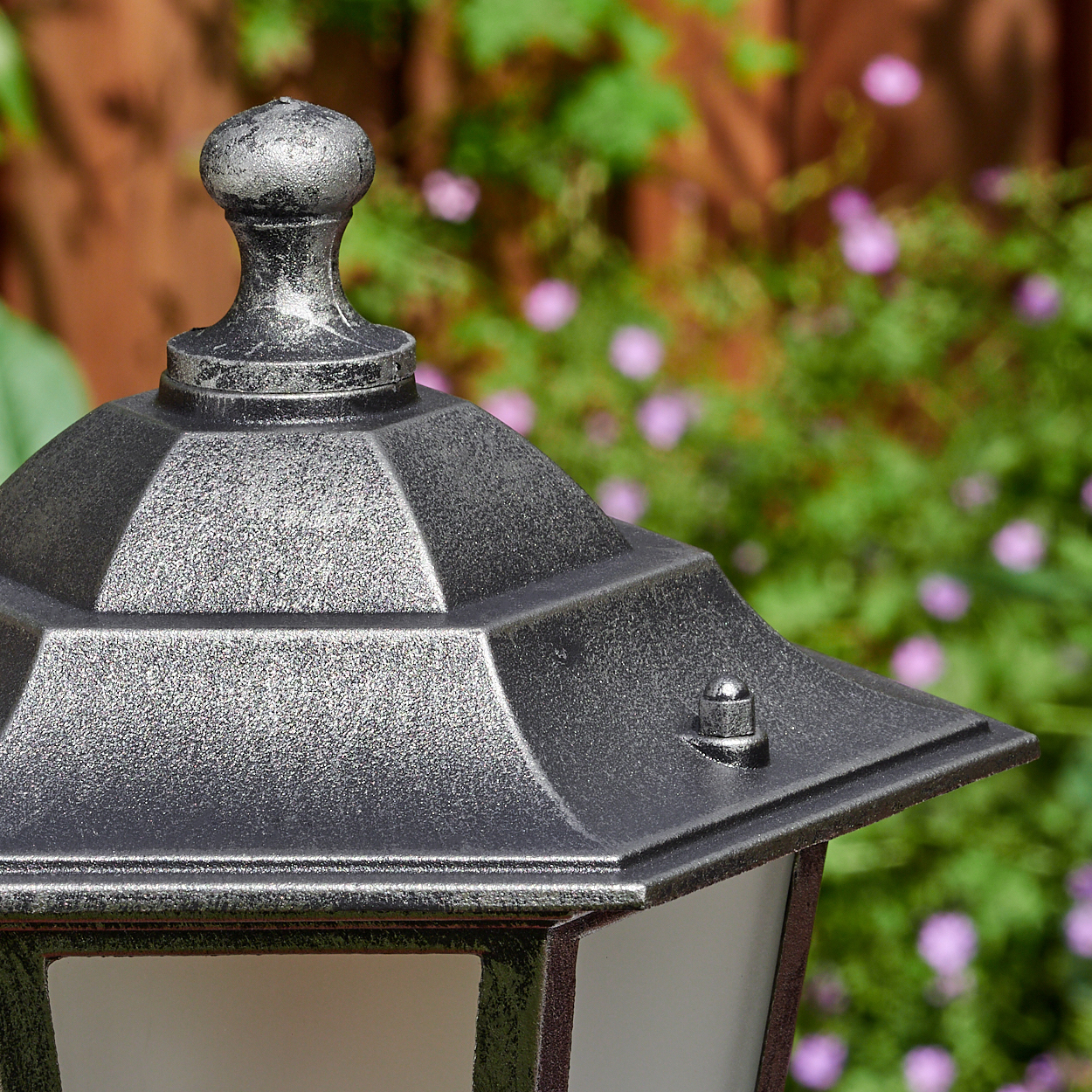 Nahaufnahme einer dekorativen Laterne mit Metallic-Finish. Im Hintergrund verschwommenes grünes Laub und rosa Blüten.