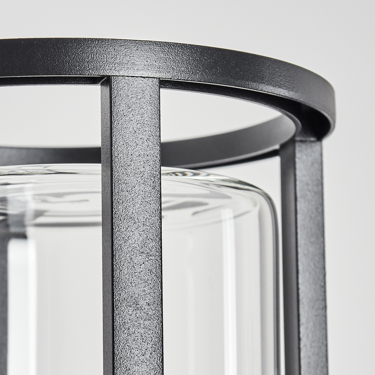 Nahaufnahme eines schwarzen Metalllaternenrahmens mit einem durchsichtigen Glaszylinder im Inneren. Der Hintergrund ist schlicht weiß.