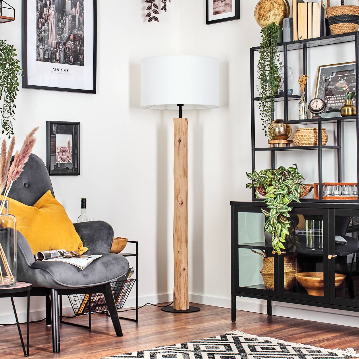 Ein Wohnzimmer mit grauem Sessel, gelbem Kissen, Holzlampe, schwarzem Regal und gemustertem Teppich.