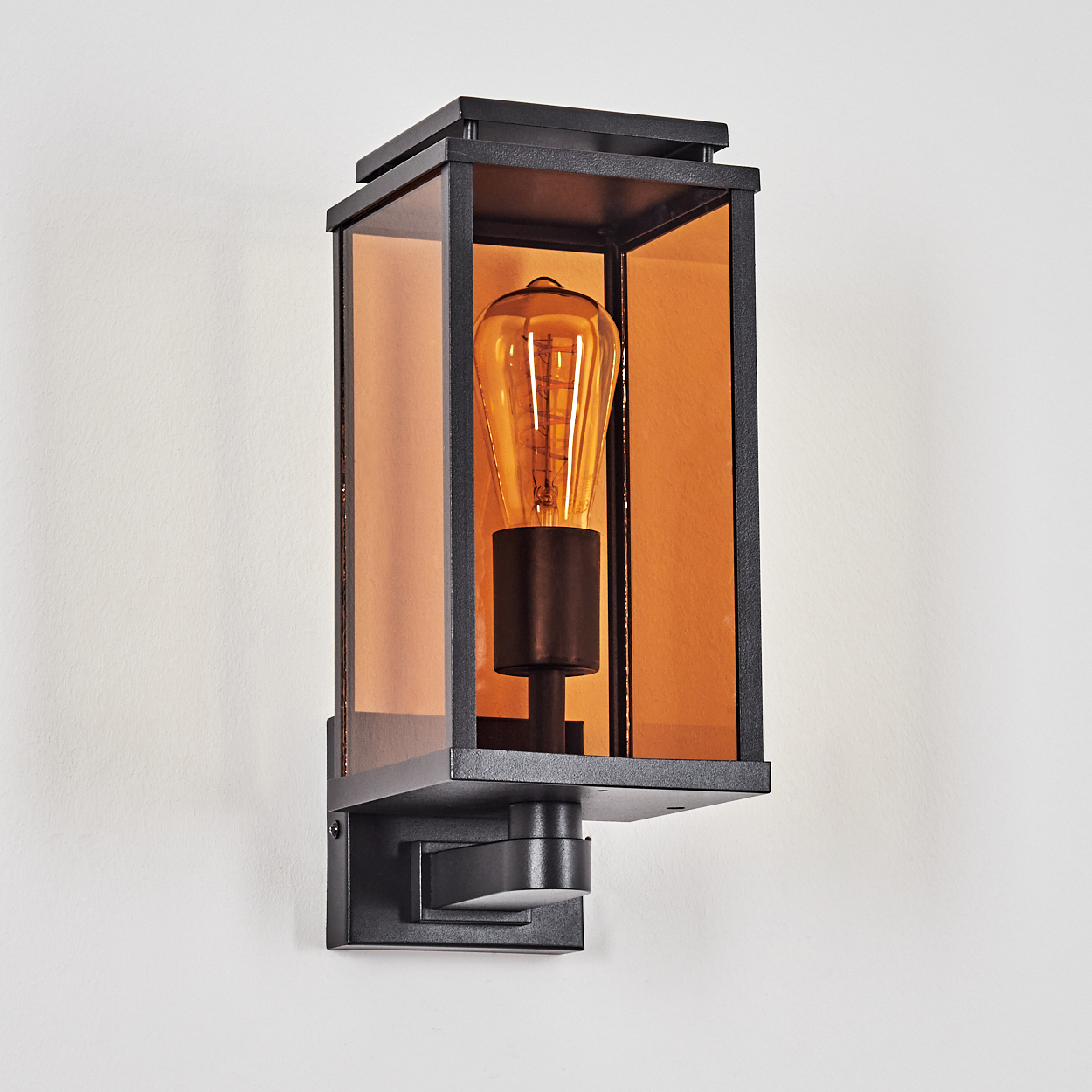 Eine schwarze, rechteckige Außenwandleuchte mit bernsteinfarbenem Glas und einer sichtbaren Glühbirne im Vintage-Stil, an einer weißen Wand montiert.