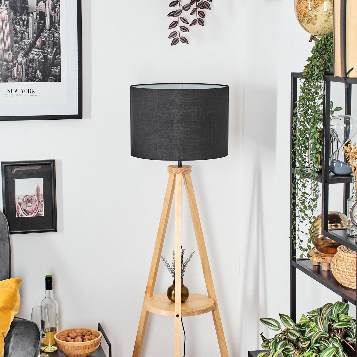 Eine hölzerne Stativlampe mit schwarzem Schirm. Ein Regal hält eine Vase. Wandkunst und ein Regal mit Pflanzen sind sichtbar.