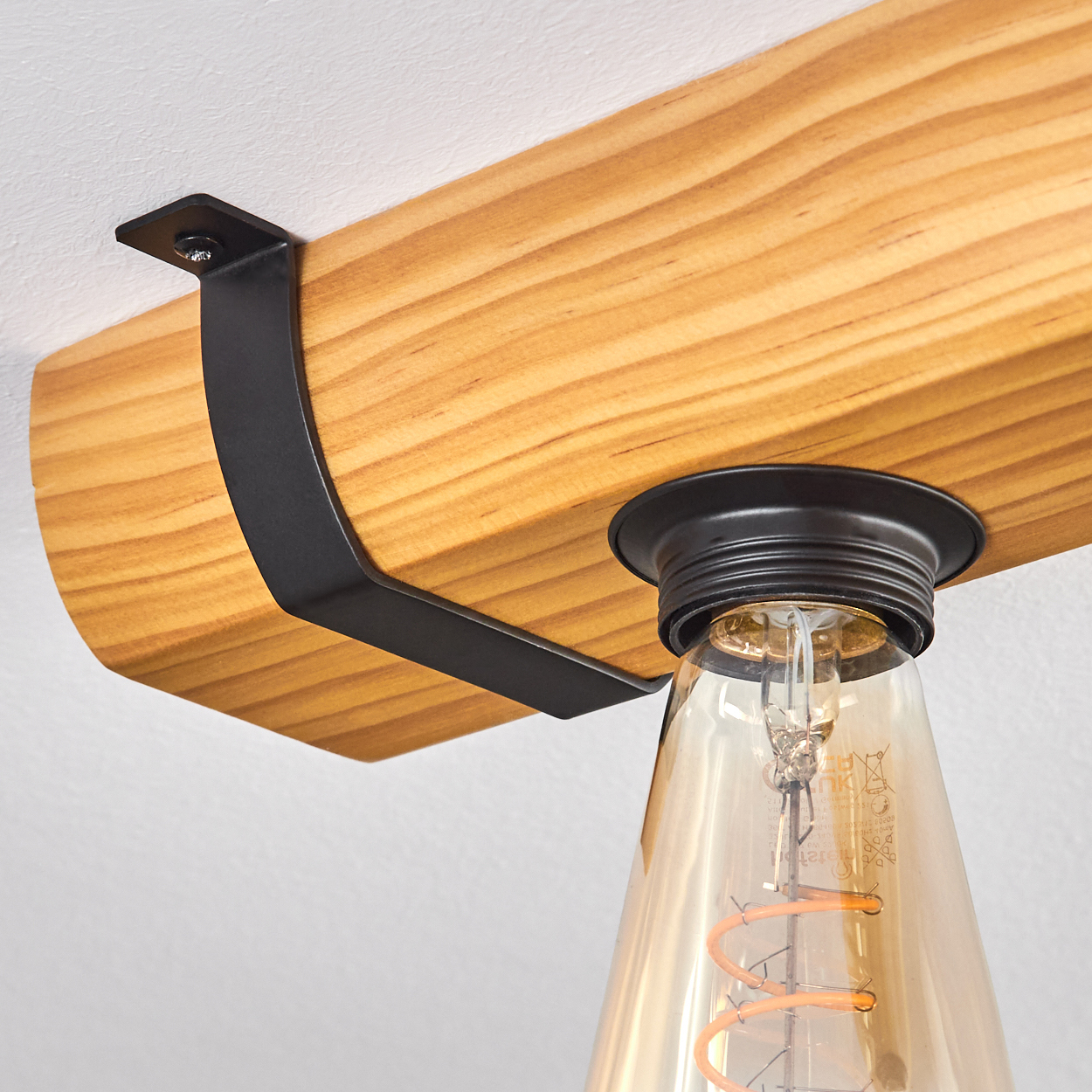 Ein Holzbalken mit schwarzem Metallbügel trägt eine Glühbirne. Die Glühbirne ist aus Glas mit sichtbarem Glühfaden.
