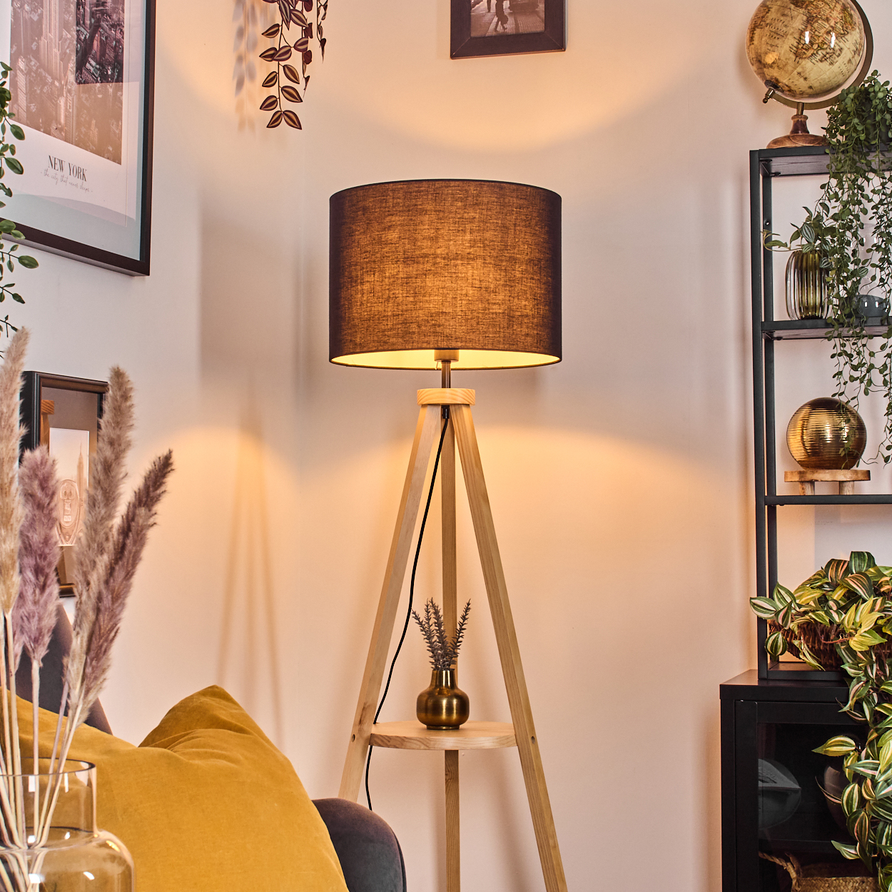 Eine Holz-Dreibein-Lampe mit schwarzem Lampenschirm. Ein Regal hält eine kleine Vase. Ein gelbes Kissen und gerahmte Kunstwerke sind im Hintergrund.