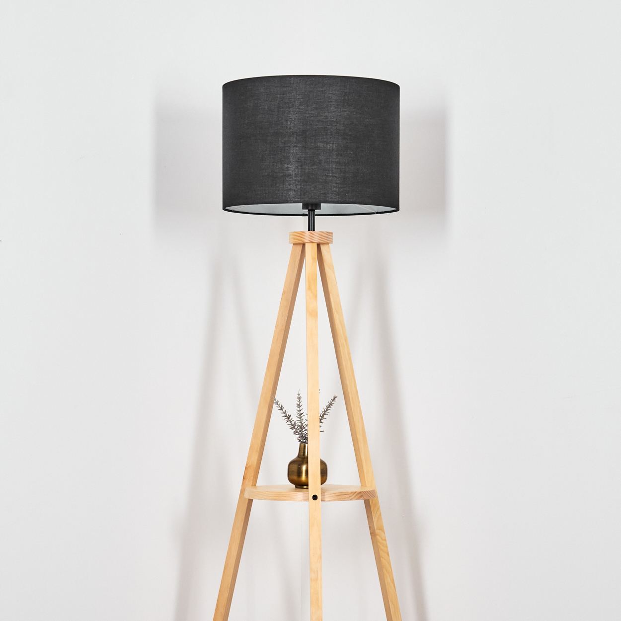 Eine Stehlampe aus Holz mit schwarzem Lampenschirm. Eine kleine Vase mit Pflanzen steht auf einem Regal. Weißer Wandhintergrund.
