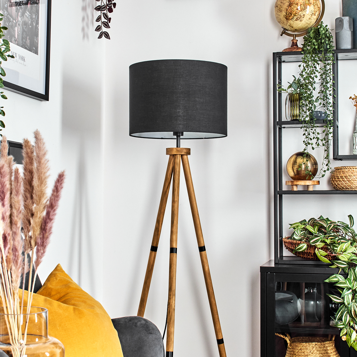 Ein Raum mit einer schwarzen Lampe, einem hölzernen Stativ und einem Regal mit Pflanzen und Dekorationen. Ein gelbes Kissen ist ebenfalls zu sehen.