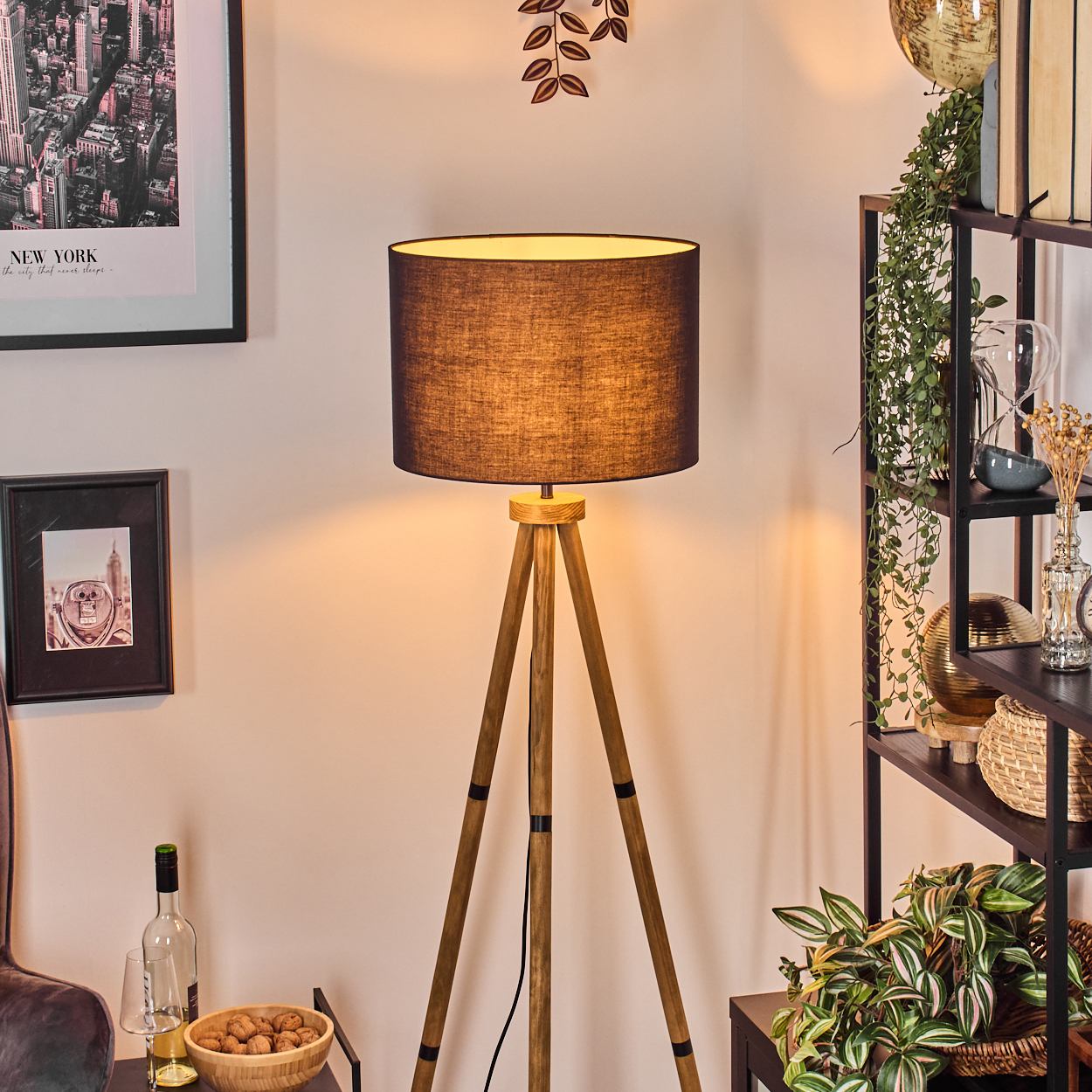 Eine Stehlampe mit schwarzem Lampenschirm. Daneben ein Regal mit Pflanzen und Dekorationen. Ein New York-Bild hängt an der Wand.