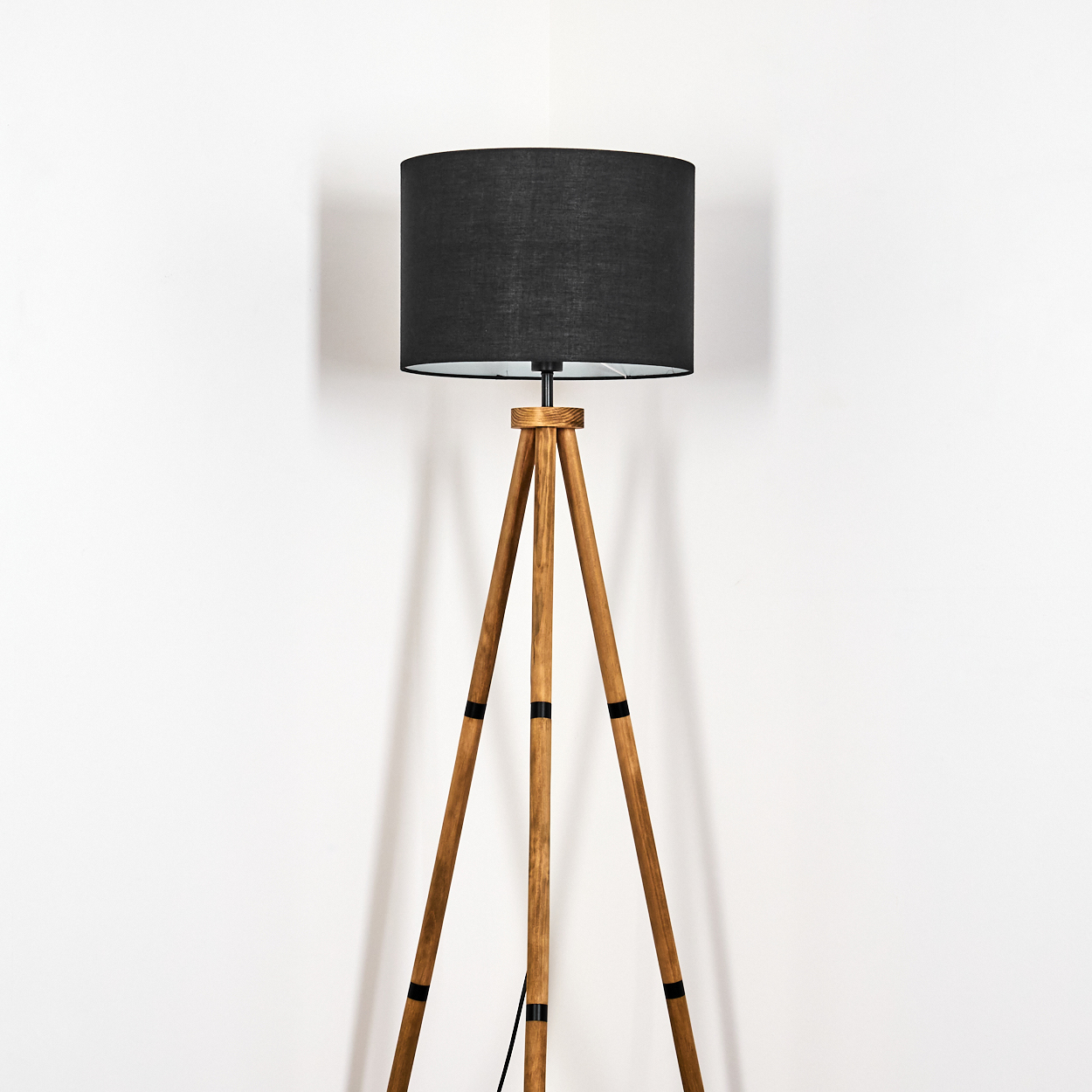 Eine hölzerne Stativ-Stehlampe mit schwarzem Lampenschirm steht vor einer weißen Wand. Die Lampe ist mittig platziert.