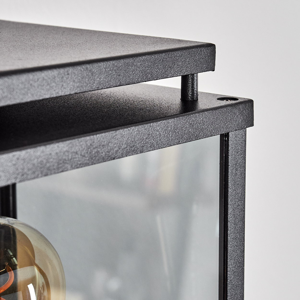 Nahaufnahme eines schwarzen, strukturierten Metallrahmens mit Glas. Im Inneren ist eine Glühbirne zu sehen. Der Hintergrund ist schlicht weiß.