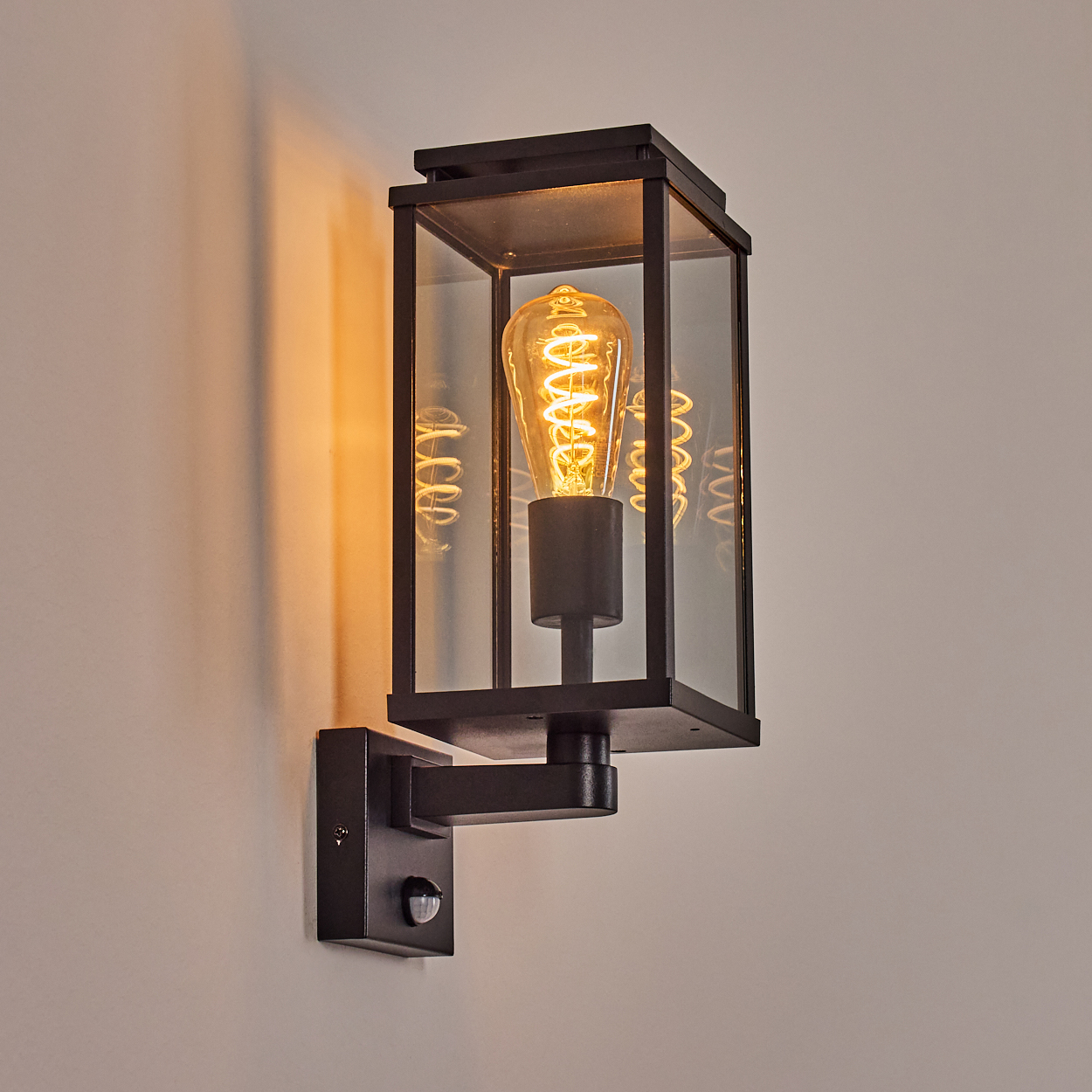 Eine schwarze Wandlampe mit Glasgehäuse, die eine leuchtende Glühbirne mit Wendel enthält. An einer hellen Wand montiert, mit Bewegungssensor.