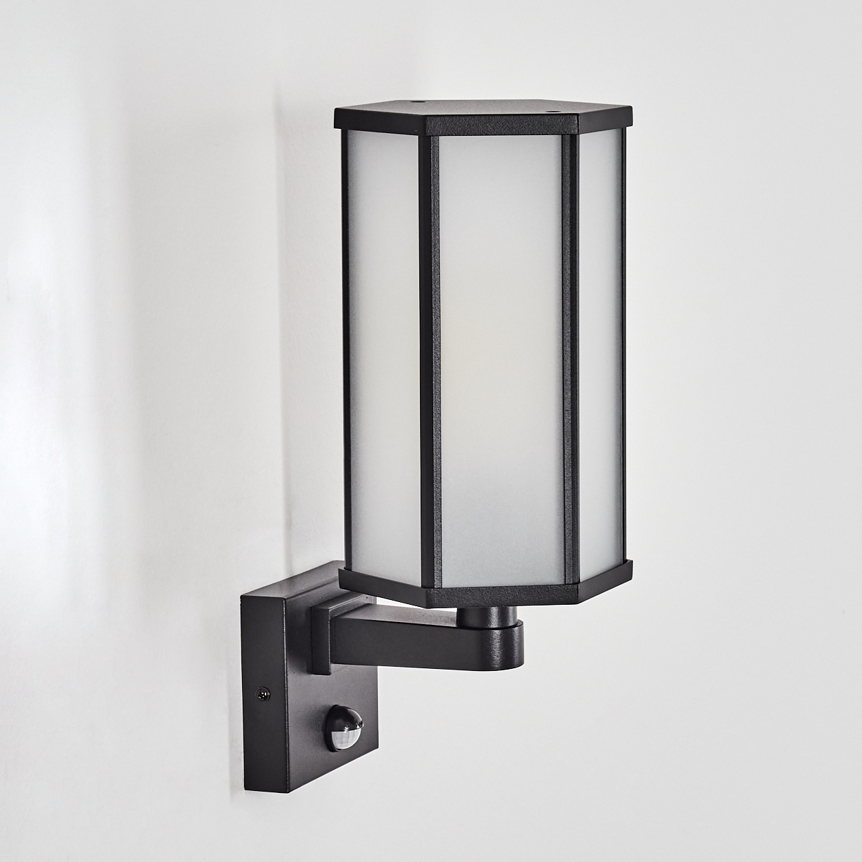 Eine schwarze Außenwandleuchte mit rechteckigem Schirm aus mattiertem Glas und Bewegungssensor, an einer weißen Wand montiert.