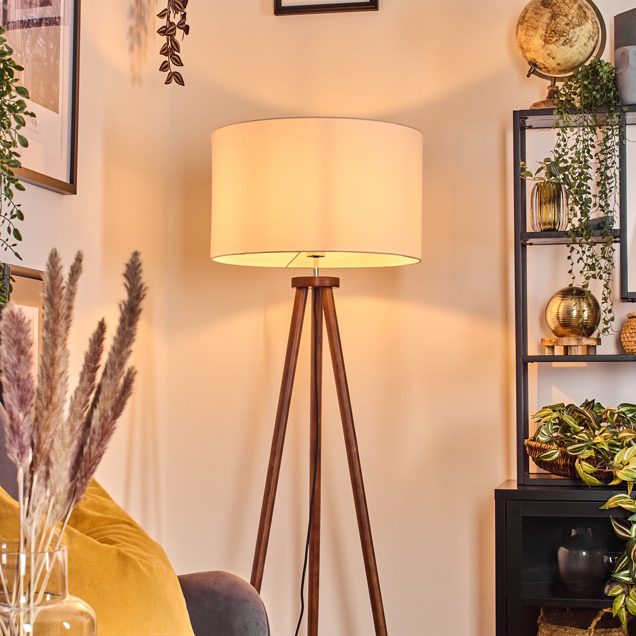Eine hölzerne Stativlampe mit weißem Lampenschirm steht in einem Raum. Regale mit Pflanzen und einem Globus sind im Hintergrund.